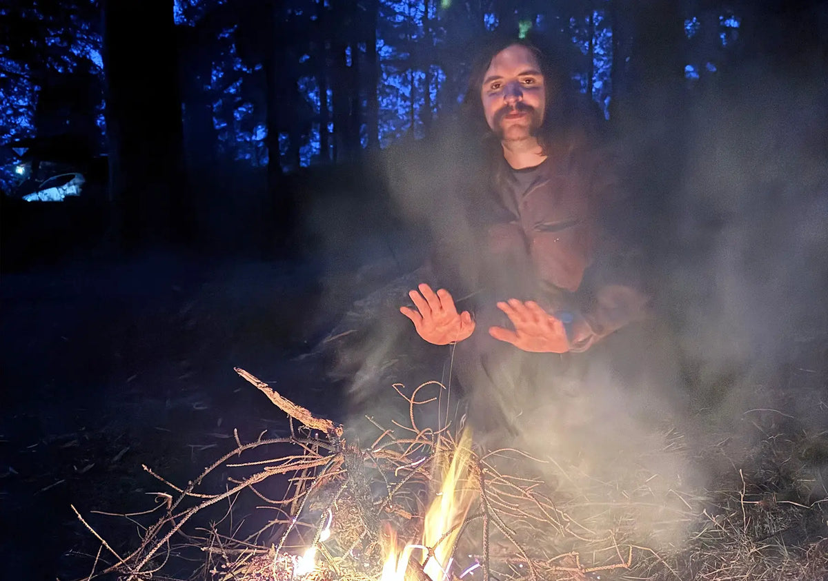 A man with long hair and a beard sits in a dark forest at night, warming himself by a small campfire with smoke rising around him.