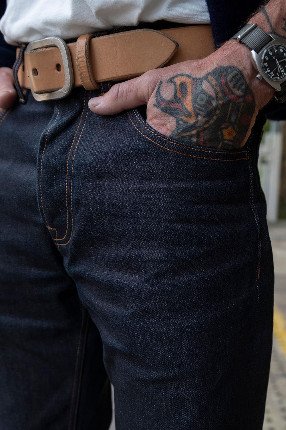 Close-up of a person wearing dark indigo denim jeans with contrast tan stitching, a tan leather belt with a silver buckle, and a white shirt tucked in. The person's tattooed hand rests on the hip pocket.