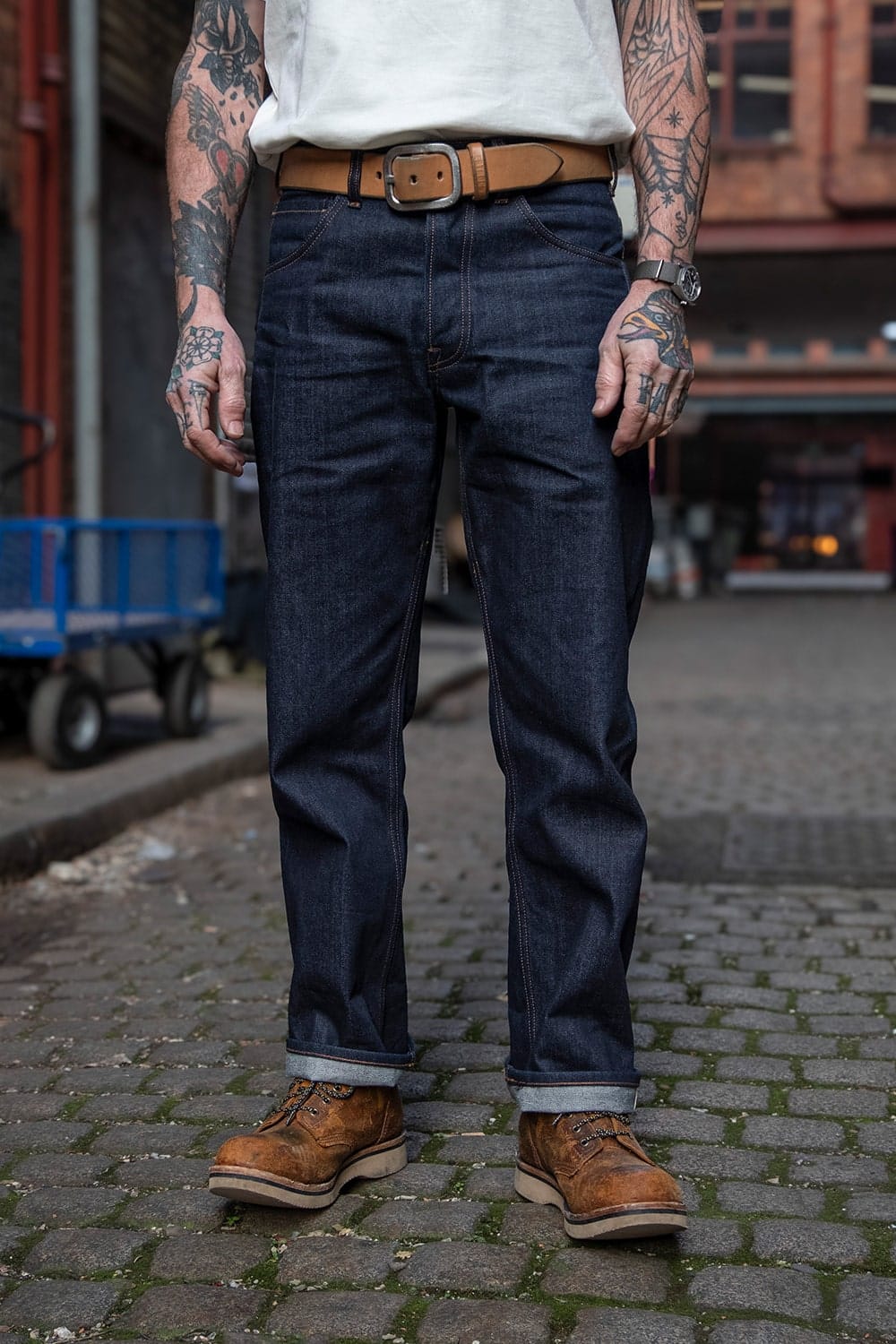 A person standing outdoors on a cobblestone street wearing dark indigo denim jeans, a white t-shirt, a brown leather belt, and brown lace-up work boots.