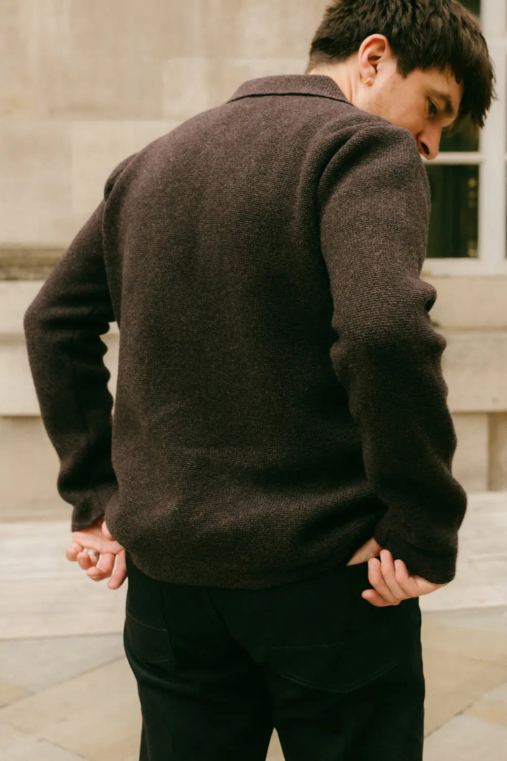 A man wearing a dark brown textured knit sweater, shown from the back, standing outdoors against a stone building background.