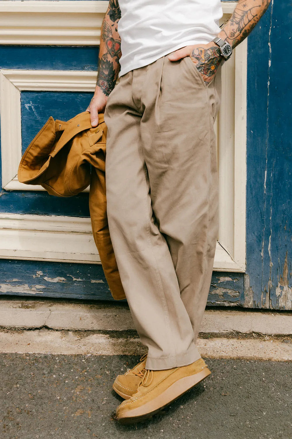 A person wearing loose beige cargo-style pants, a white t-shirt, and tan suede shoes, leaning against a blue wooden door and holding a brown jacket.
