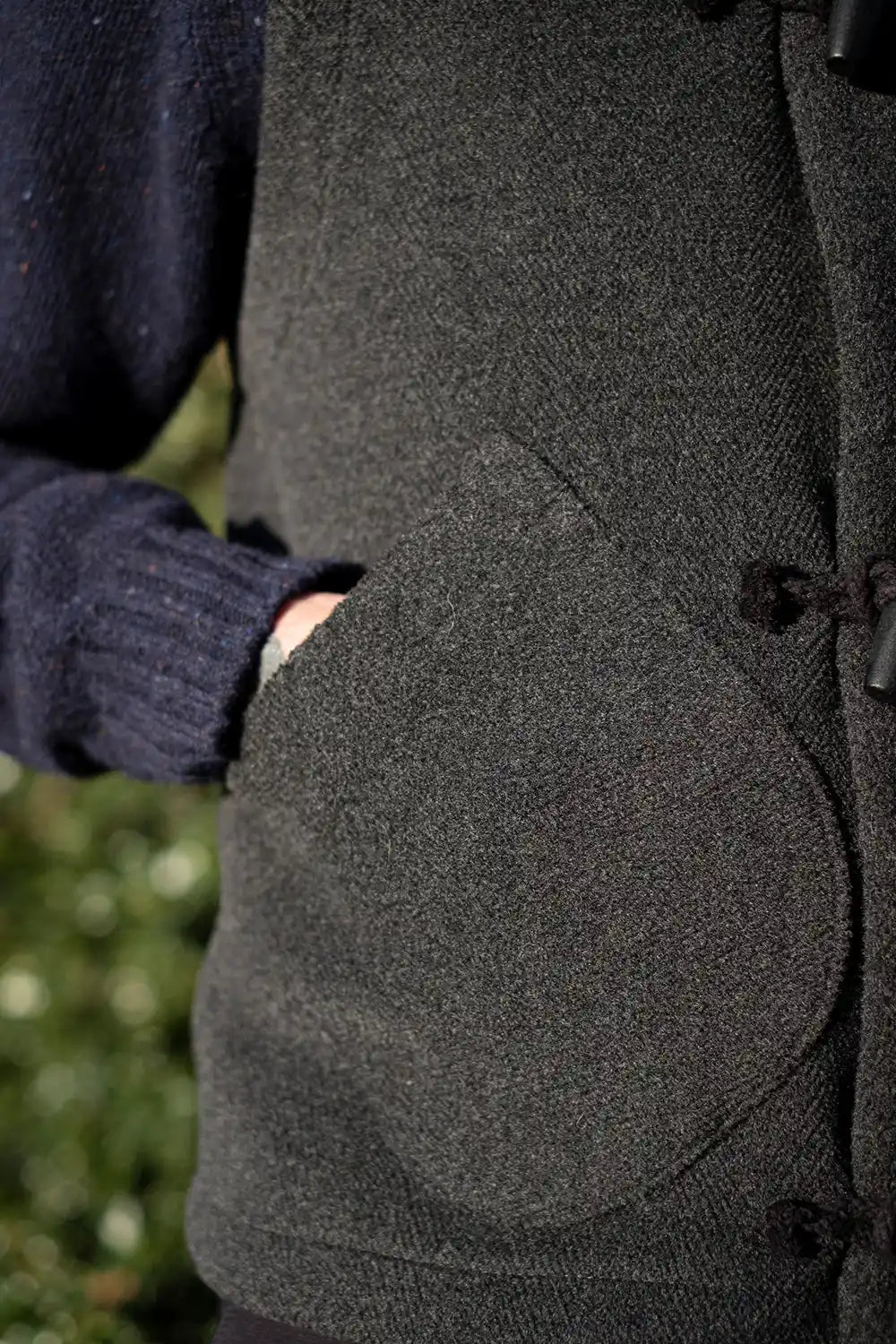 Close-up of a person wearing a dark charcoal gray wool vest with a large front patch pocket and toggle closures, over a navy blue knit sweater.