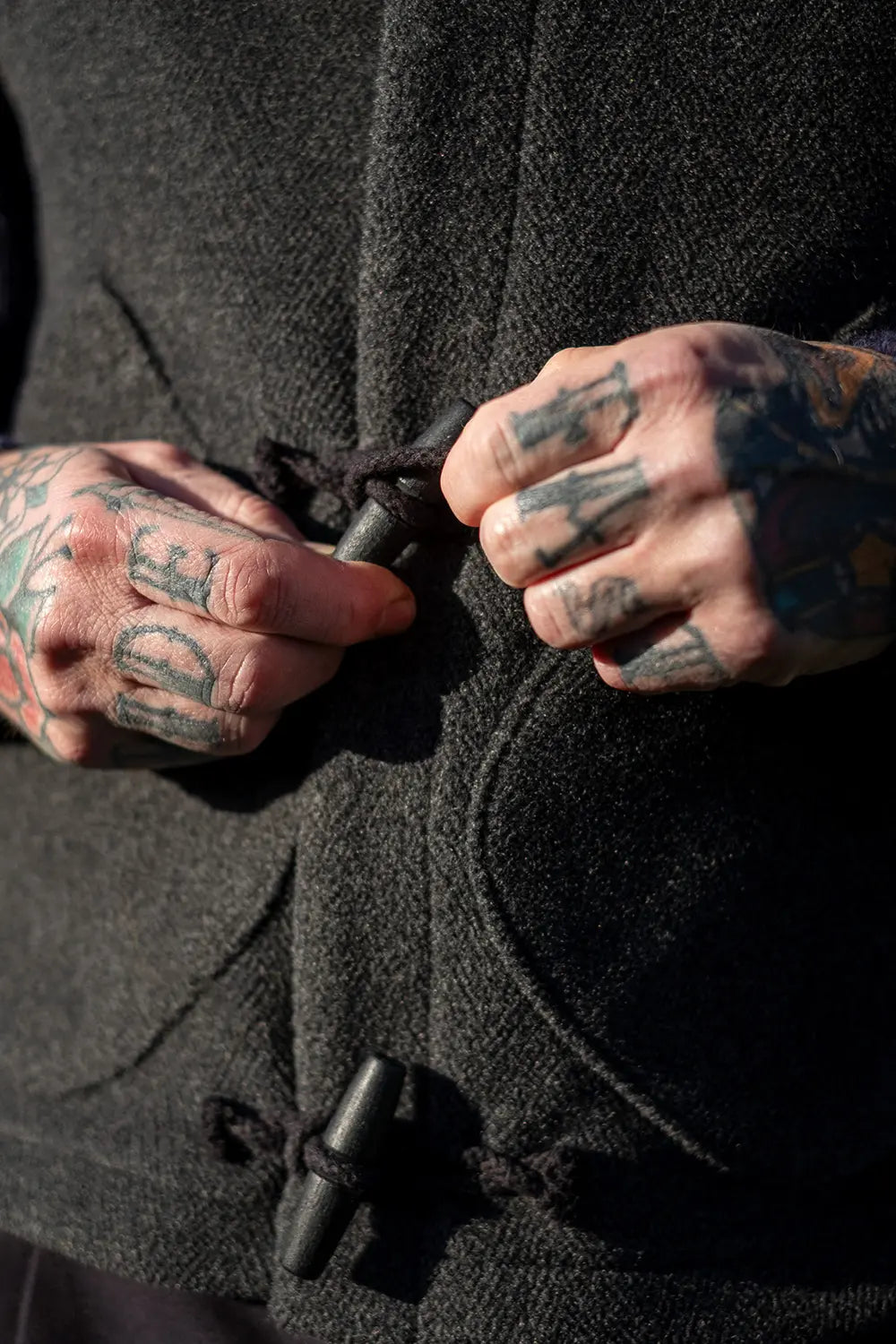 Close-up of a person's tattooed hands adjusting the toggle closure of a dark charcoal wool vest.