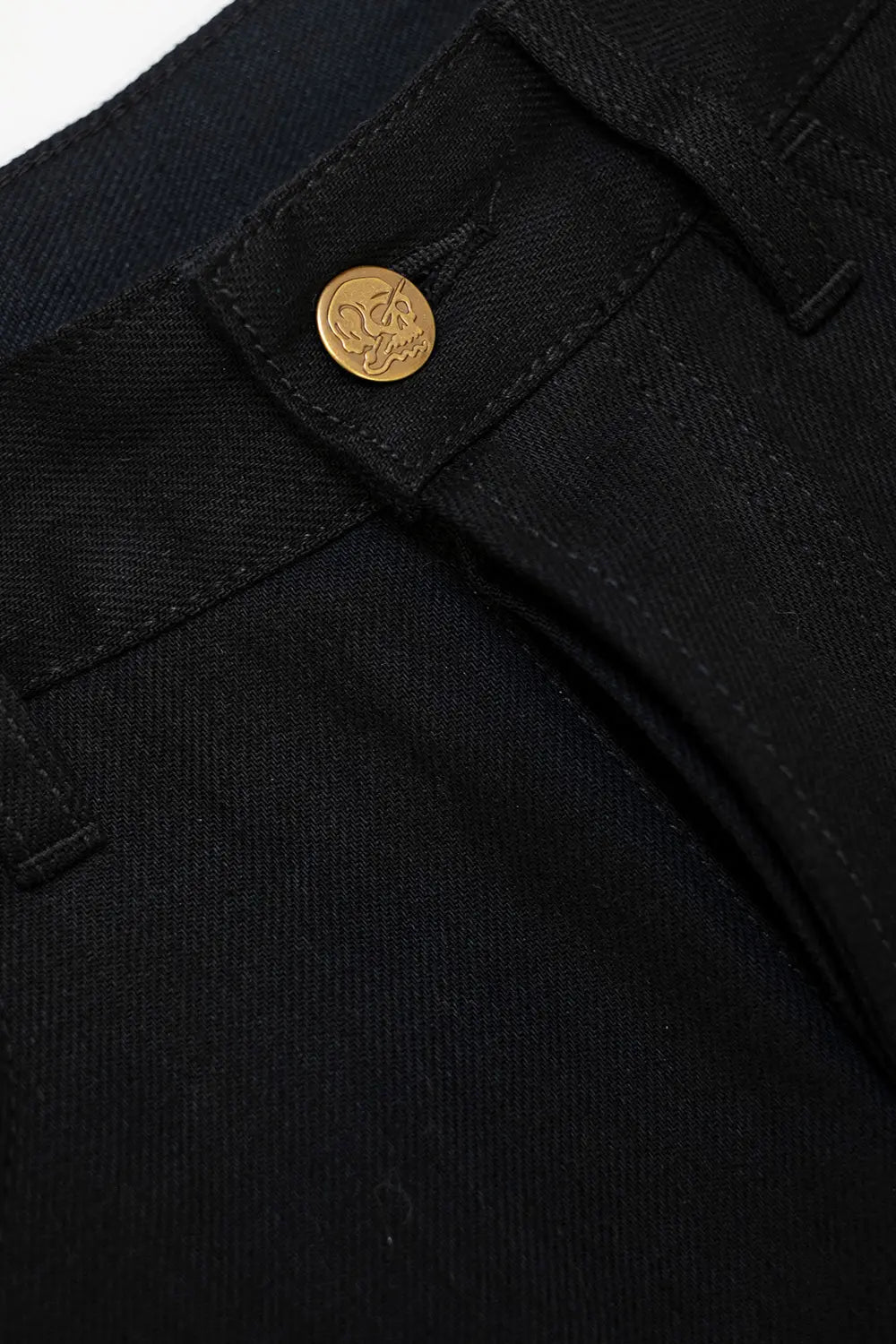 Close-up of black denim pants showing the waistband, belt loops, and a round gold-tone button with an embossed logo.