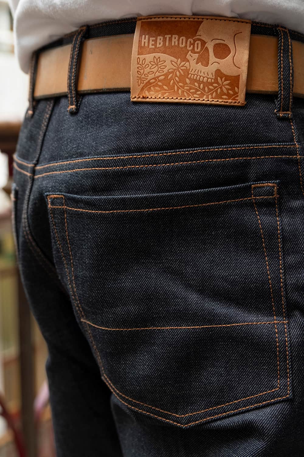 Close-up of the back of dark indigo denim jeans with contrast tan stitching, a tan leather belt, and a brown leather patch on the waistband featuring a skull design and the brand name HEBTROCO.