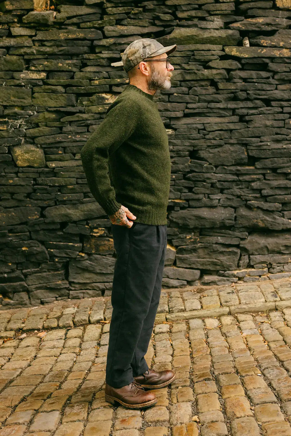 A man wearing an olive green knit wool jumper, dark trousers, brown leather shoes, and a camouflage cap stands in profile against a dark stone wall on a cobblestone path.