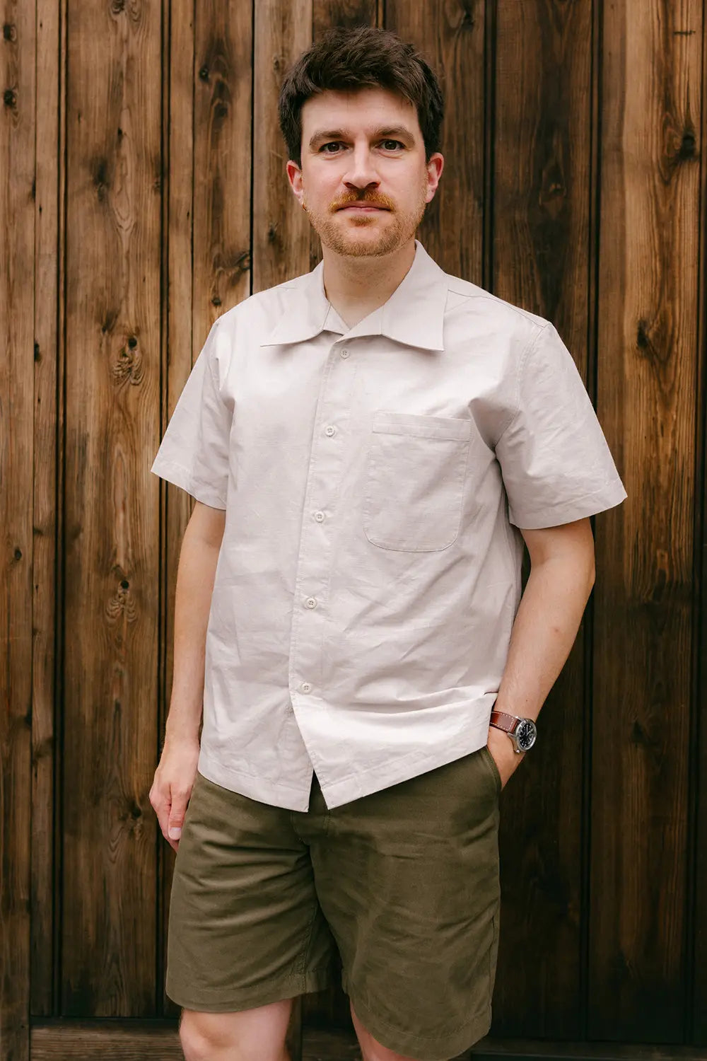 A man wearing a light beige short-sleeve button-up shirt and olive green shorts, standing against a rustic brown wooden plank wall.