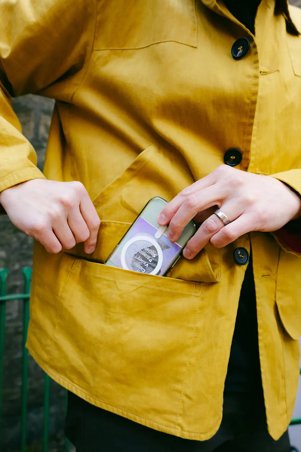 A close-up of a person wearing a mustard yellow waxed cotton utility jacket, showing their hands reaching into a front pocket to retrieve a smartphone.