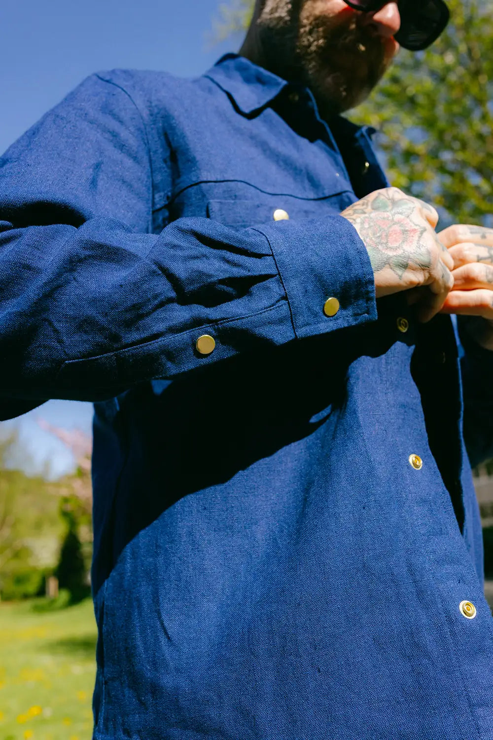 A close-up of a man wearing a dark indigo blue long-sleeve button-up shirt with brass buttons, outdoors under bright sunlight.