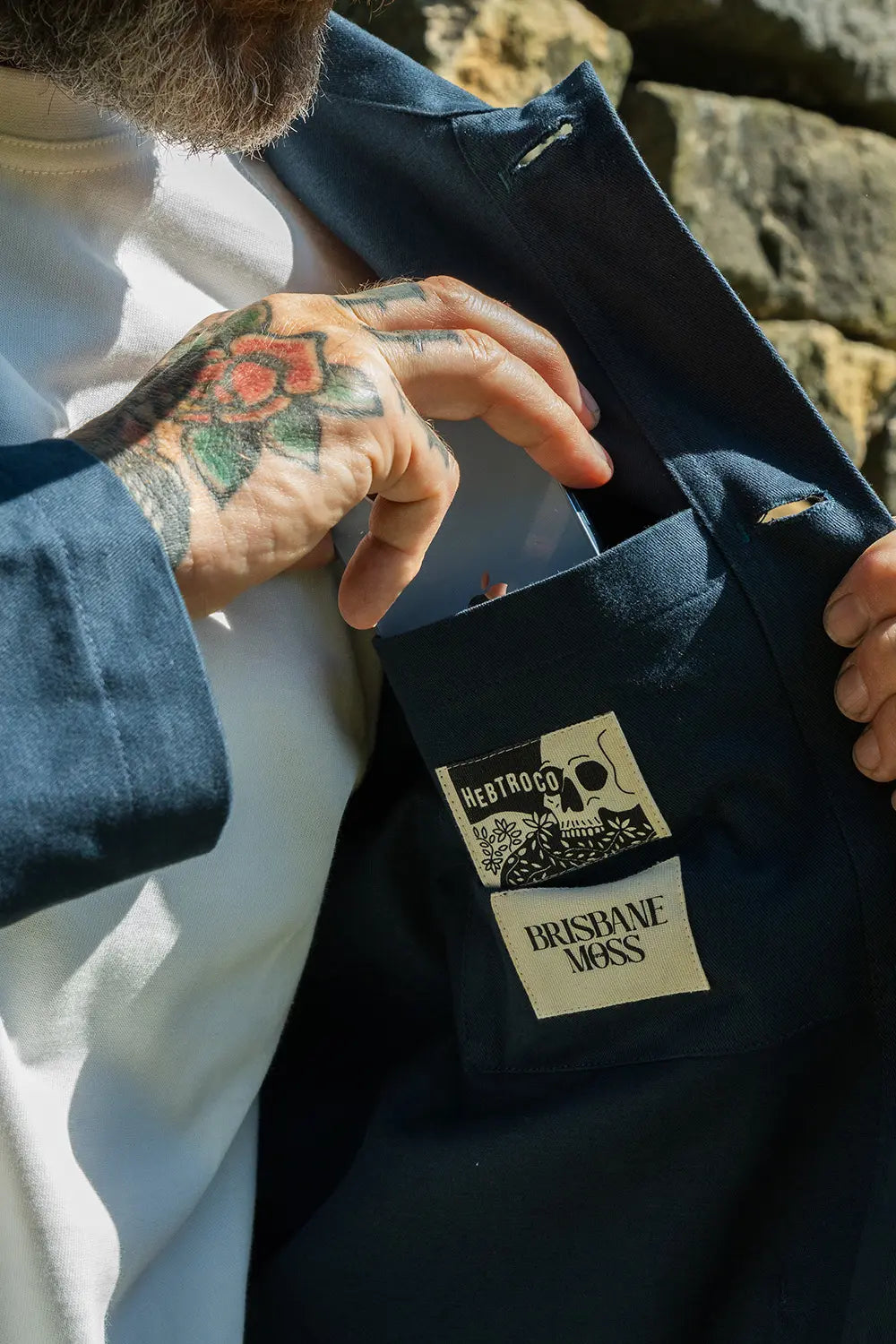 A close-up of a person wearing a dark navy blue mistle jacket over a white t shirt, with their tattooed hand reaching into the pocket to show a sewn-on brand patch that reads 'BRISHE MOSS' and 'HERITAGE'.