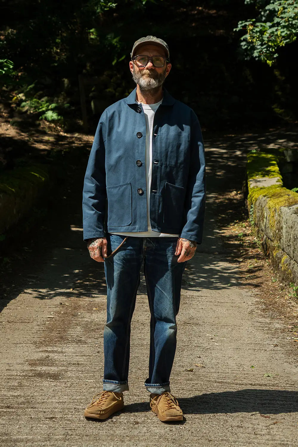 A man wearing a blue mistle jacket, blue jeans, and brown suede boots stands on a sunlit gravel path in a wooded outdoor setting.