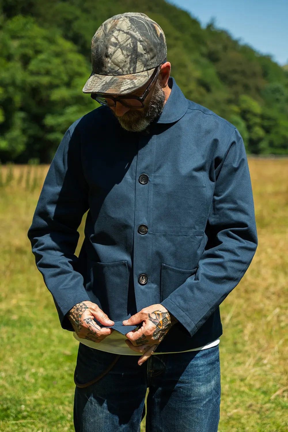 A man wearing a navy blue button-up work jacket, blue jeans, and a camouflage baseball cap stands in a grassy field with trees in the background.