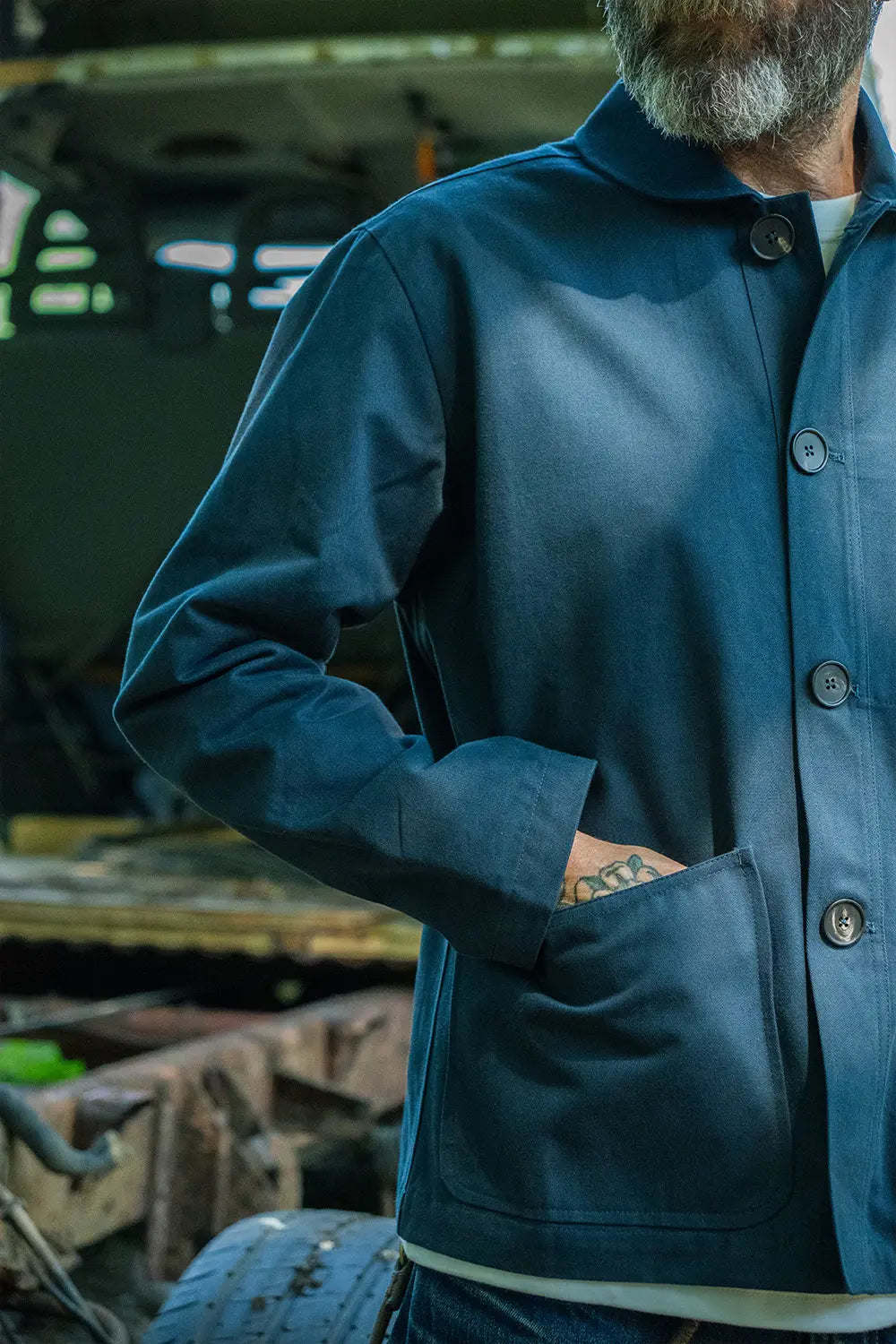 A close-up of a man wearing a dark blue button-up mistle jacket with a collar and front patch pocket, standing in an industrial setting.
