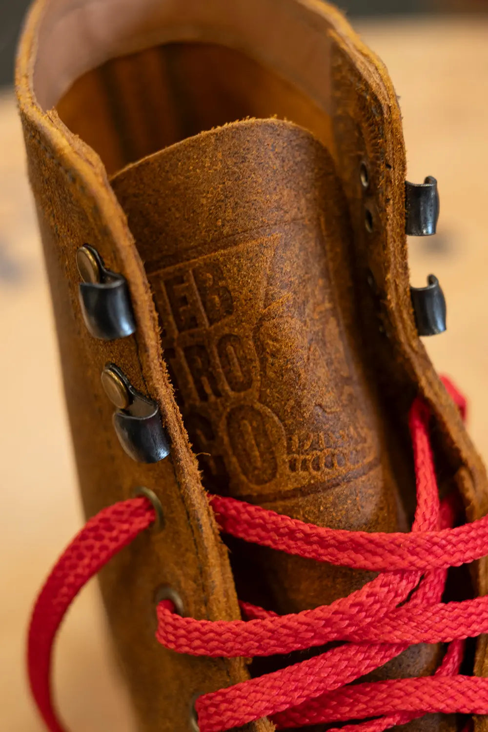 Close-up of a brown leather lace-up boot with bright red laces and black metal eyelets, showing embossed branding on the inner collar.