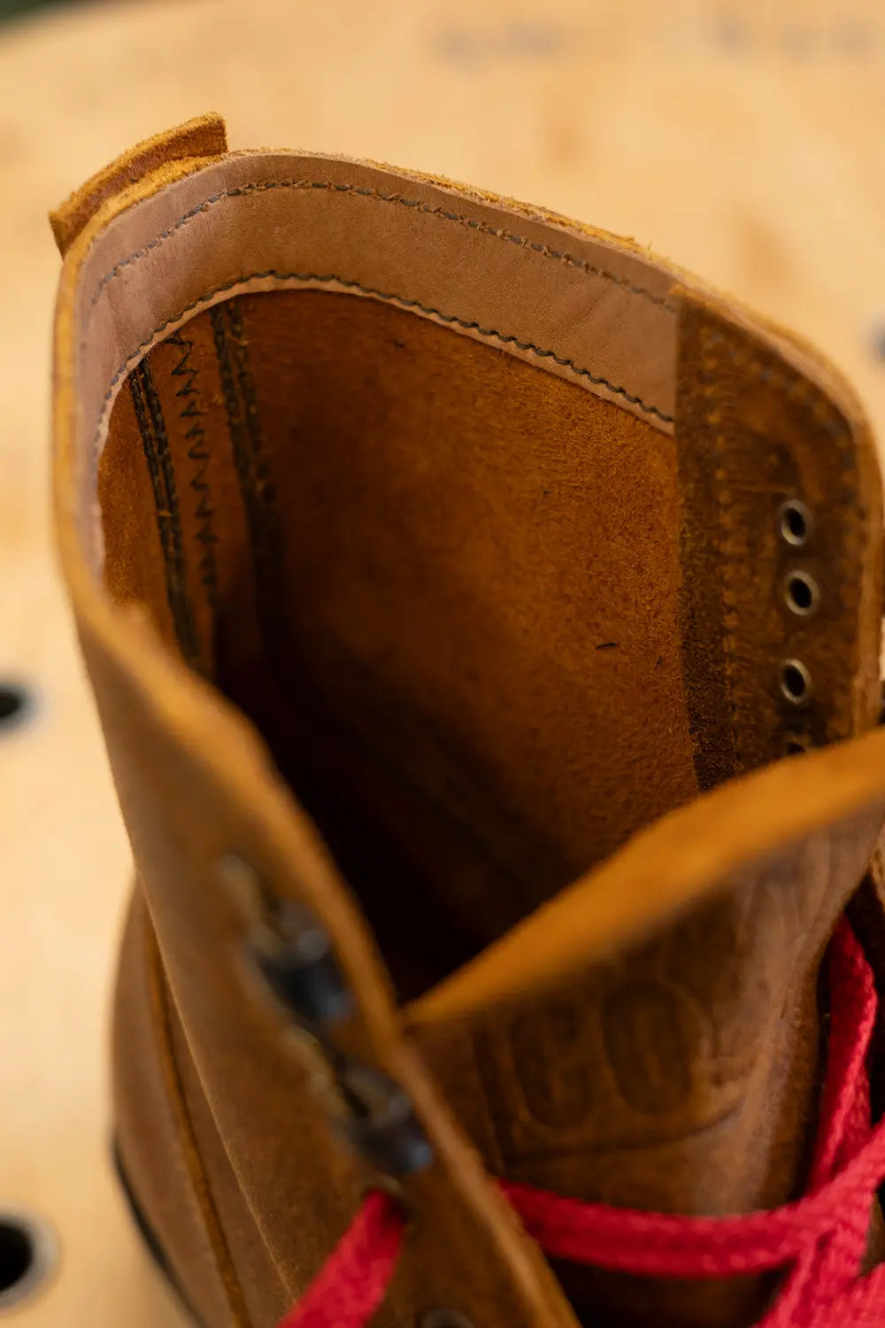 Close-up of a brown leather lace-up boot showing the interior lining, stitching, eyelets, and bright red laces against a blurred tan background.