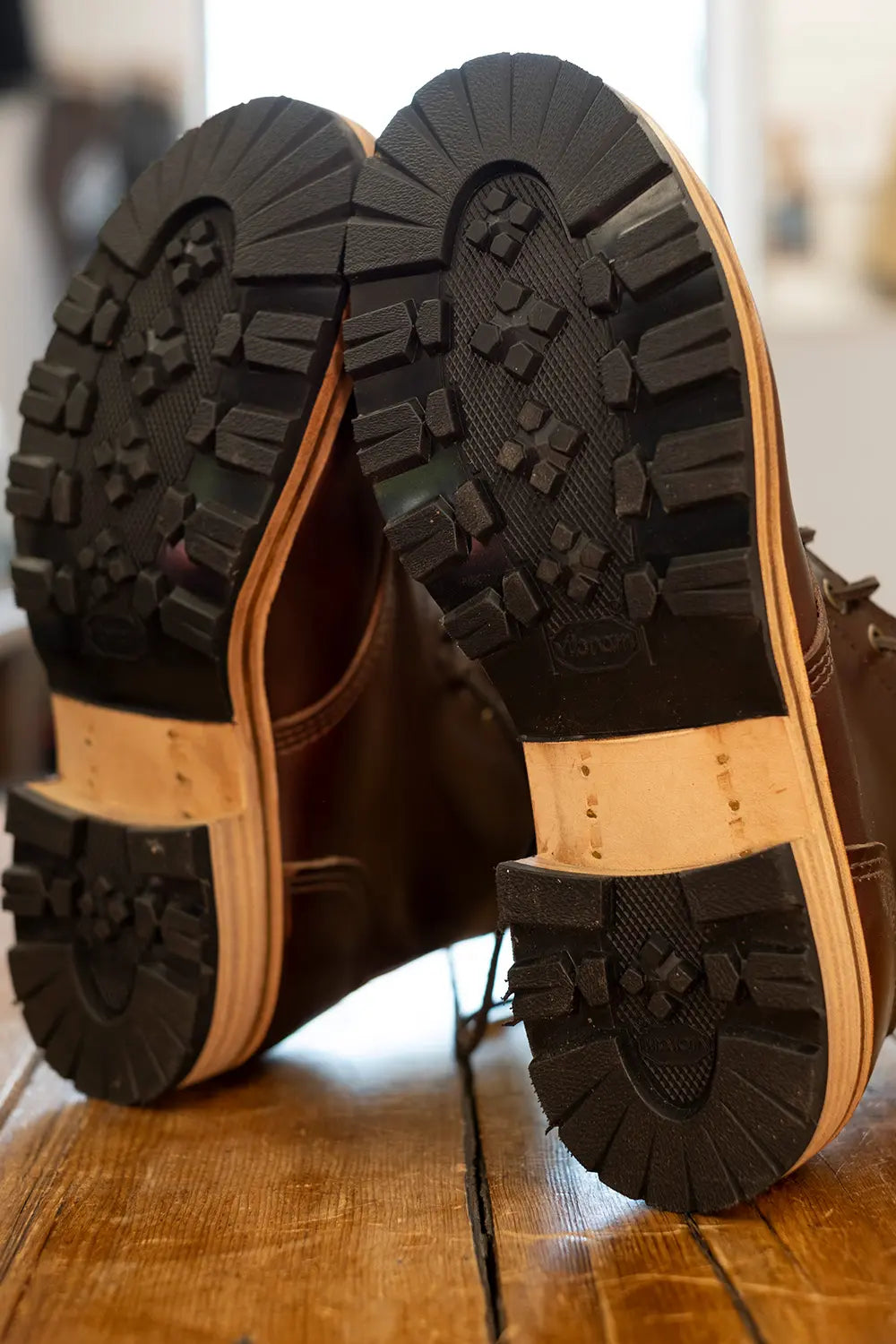 Close-up of the soles of a pair of dark brown leather boots resting on a wooden floor, showing the tread pattern and tan midsoles.