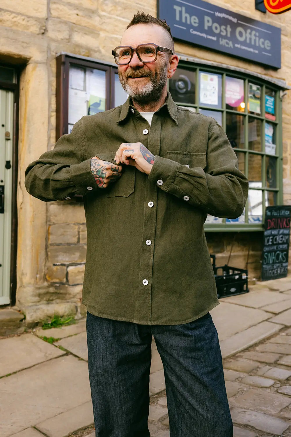 A man wearing an olive green button-up shirt and dark blue jeans stands in front of a stone building with a 'The Post Office' sign.