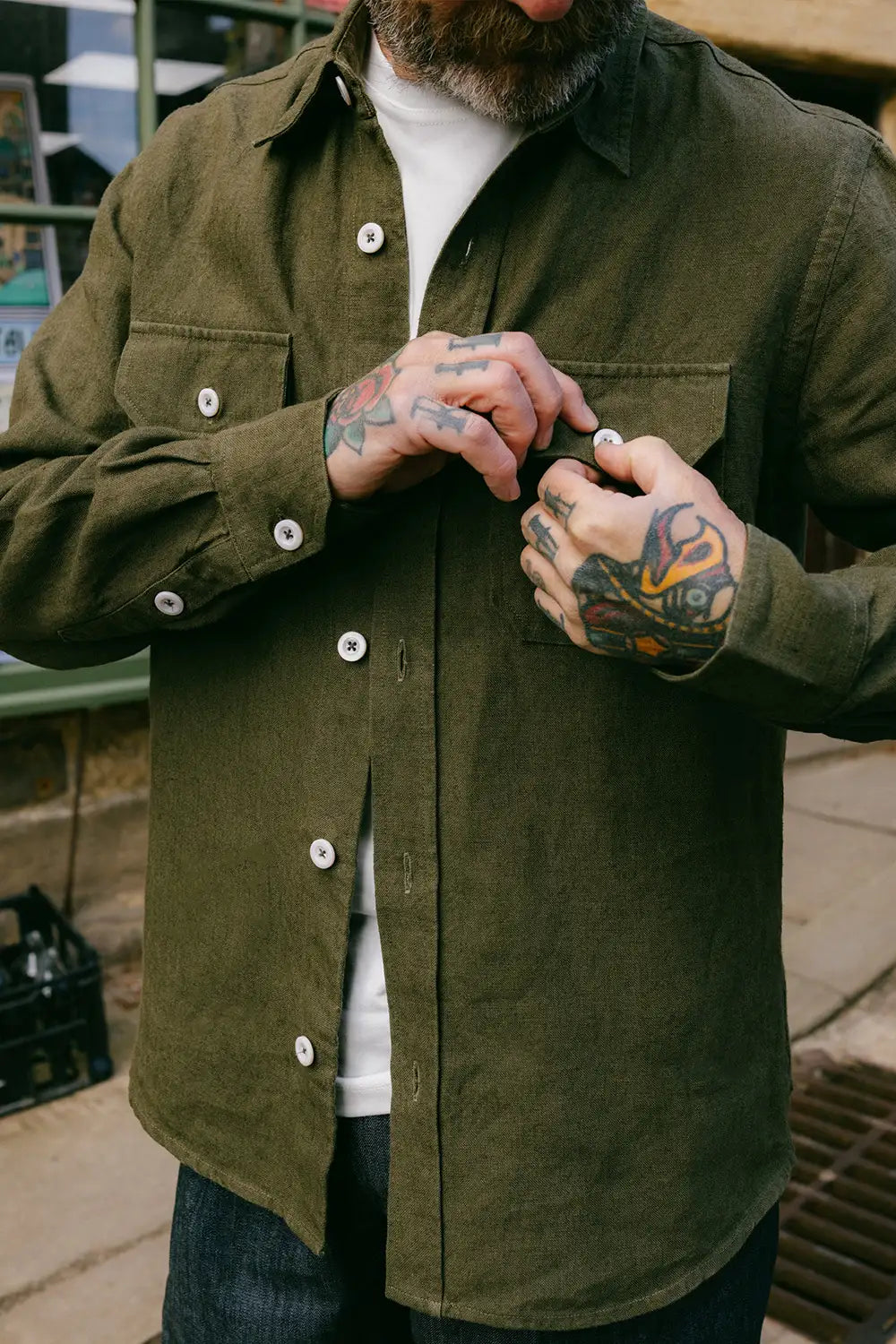 A man wearing an olive green button-up overshirt over a white t-shirt and dark jeans, with his hands adjusting the collar and buttons. The background is a slightly blurred outdoor urban setting.