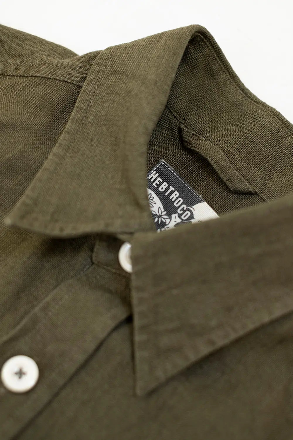 Close-up of a dark olive green button-down shirt collar with white buttons and a visible neck label reading 'HEBTROCO' with a flower logo.