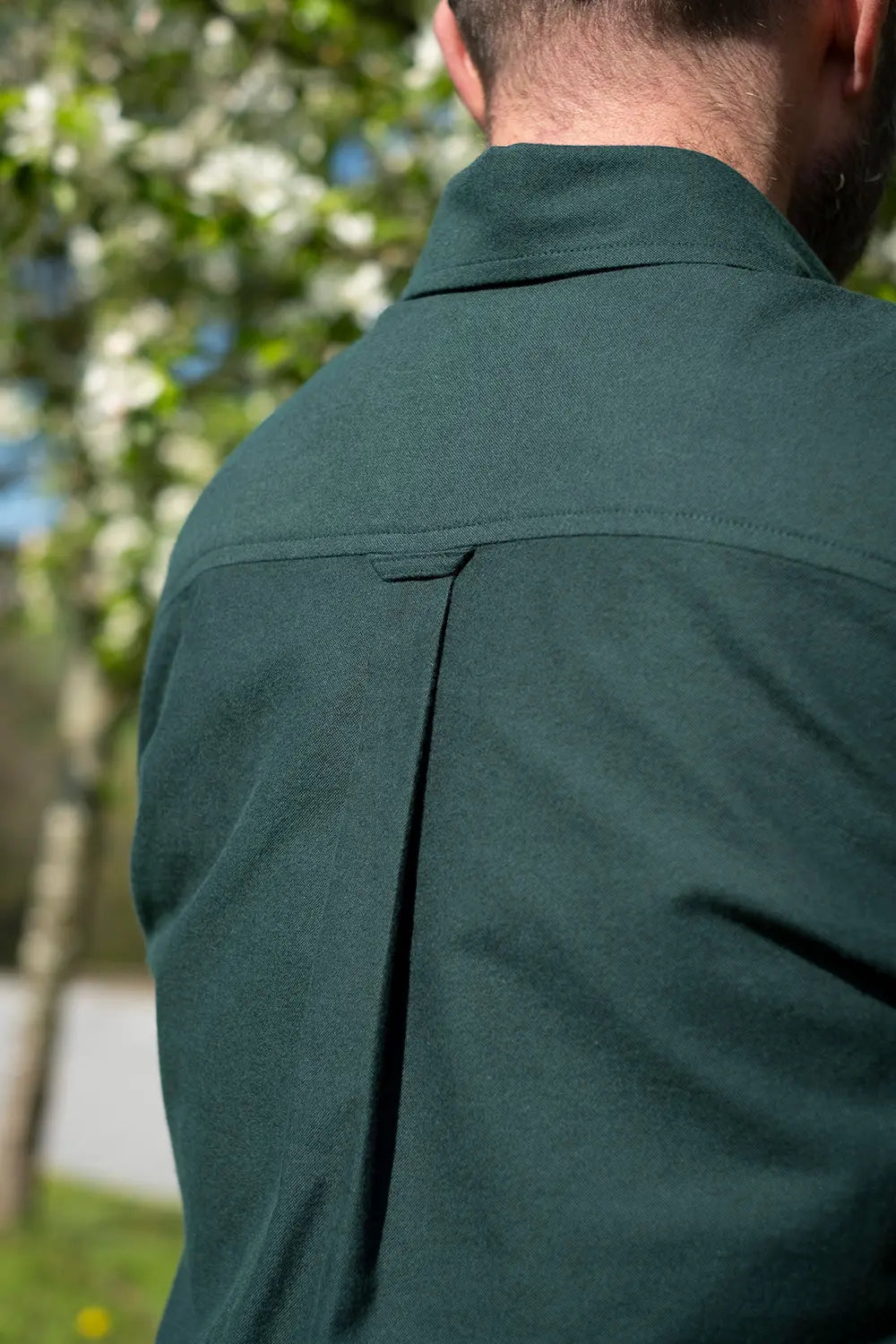 Close-up back view of a man wearing a dark green collared shirt outdoors with blurred trees in the background.