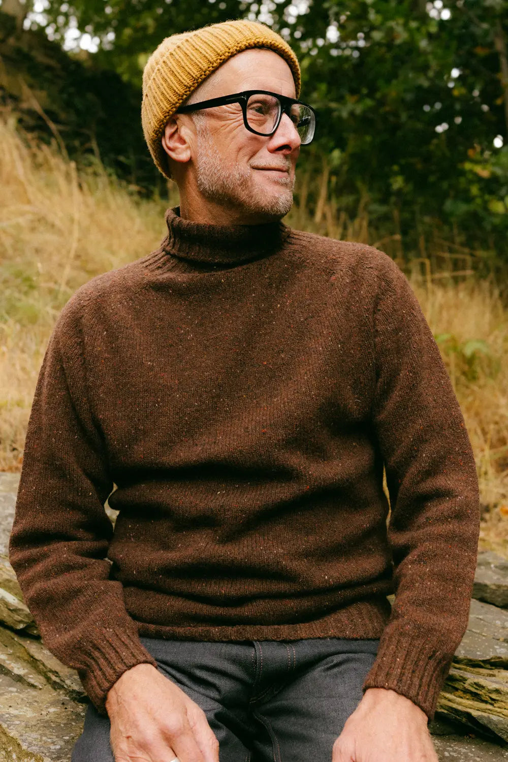 A man wearing a brown knit turtleneck sweater, a mustard yellow beanie, and black-rimmed glasses, sitting outdoors on a stone wall with dry grass and greenery in the background.