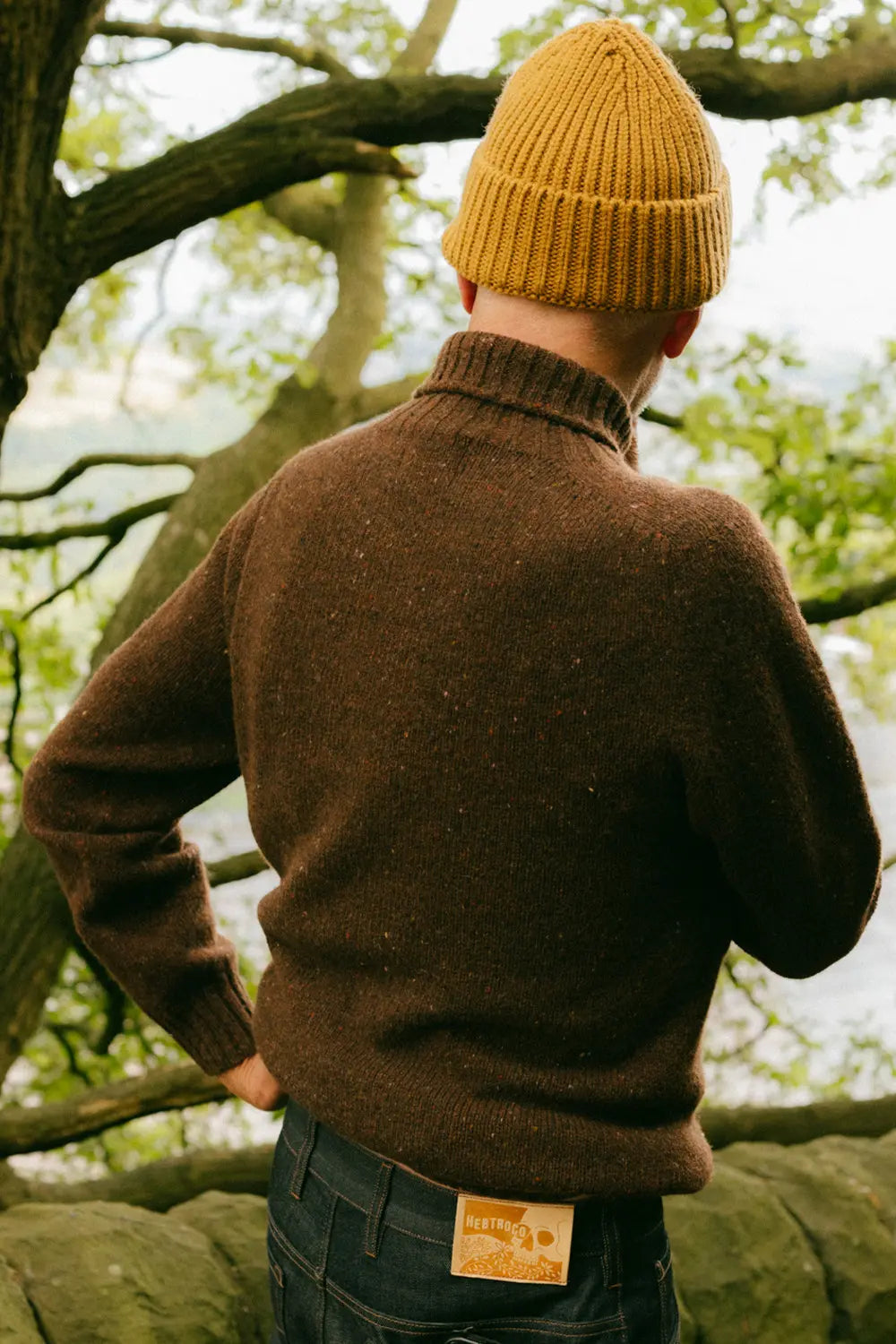 A person seen from behind wearing a brown knit sweater, dark blue jeans, and a mustard yellow beanie, standing outdoors in front of trees and rocks.