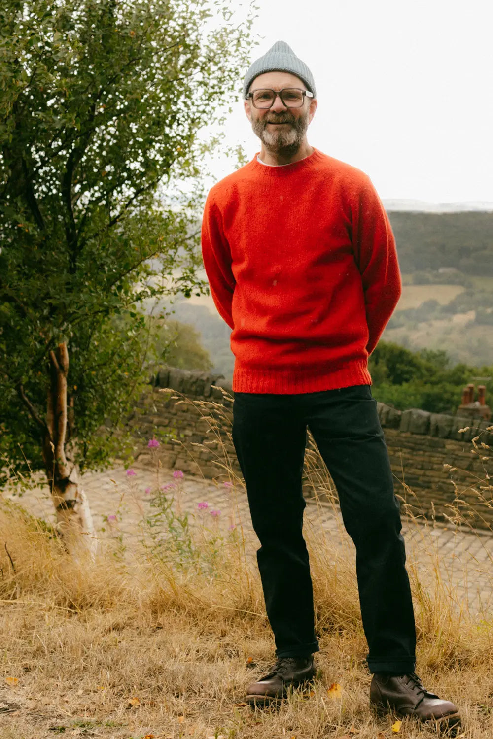 A man wearing a bright red knit jumper, black trousers, brown shoes, and a gray beanie stands outdoors in a grassy field with trees and hills in the background.