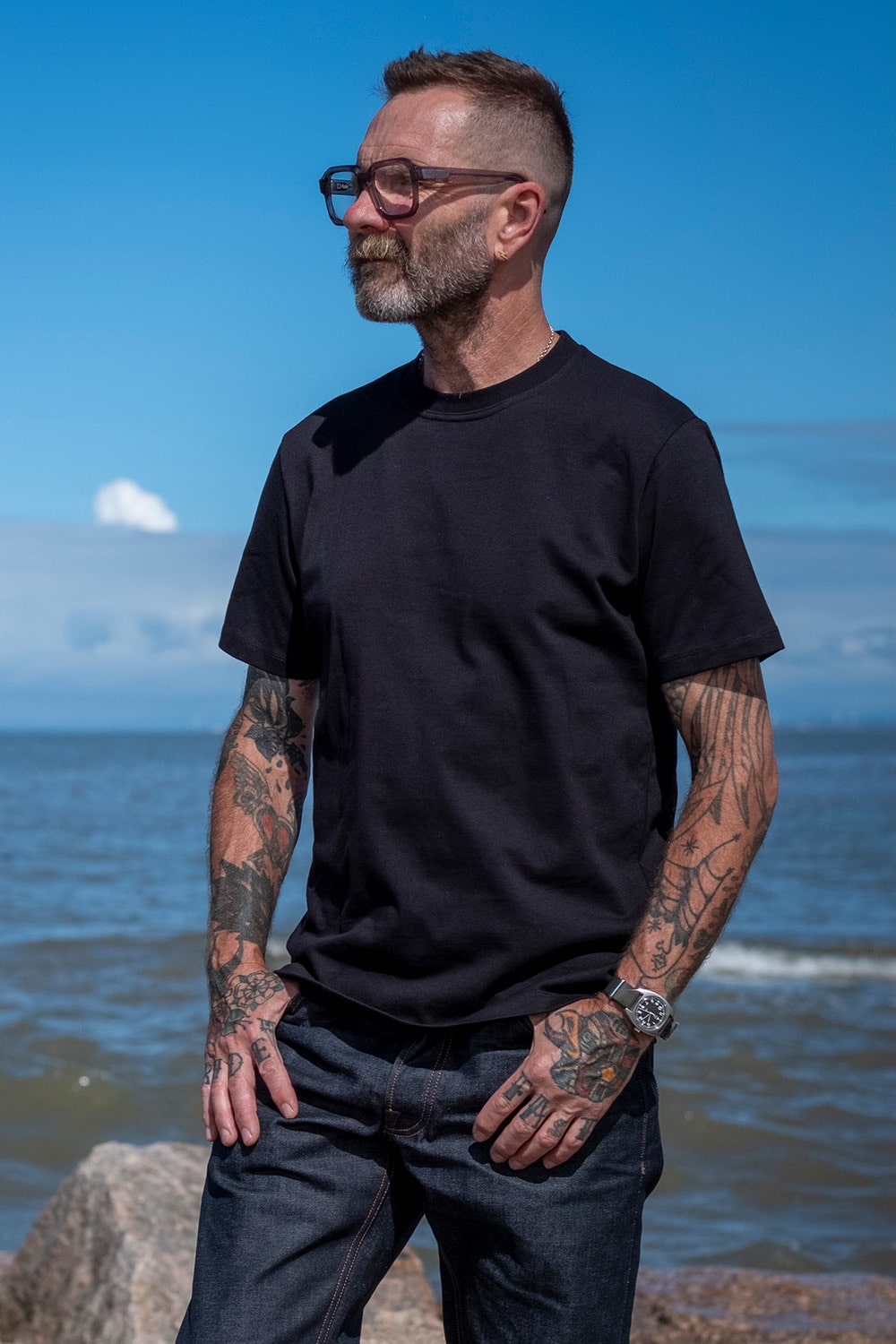 A man with tattoos and glasses stands outdoors by the water wearing a plain black short-sleeve crewneck t-shirt and dark jeans.