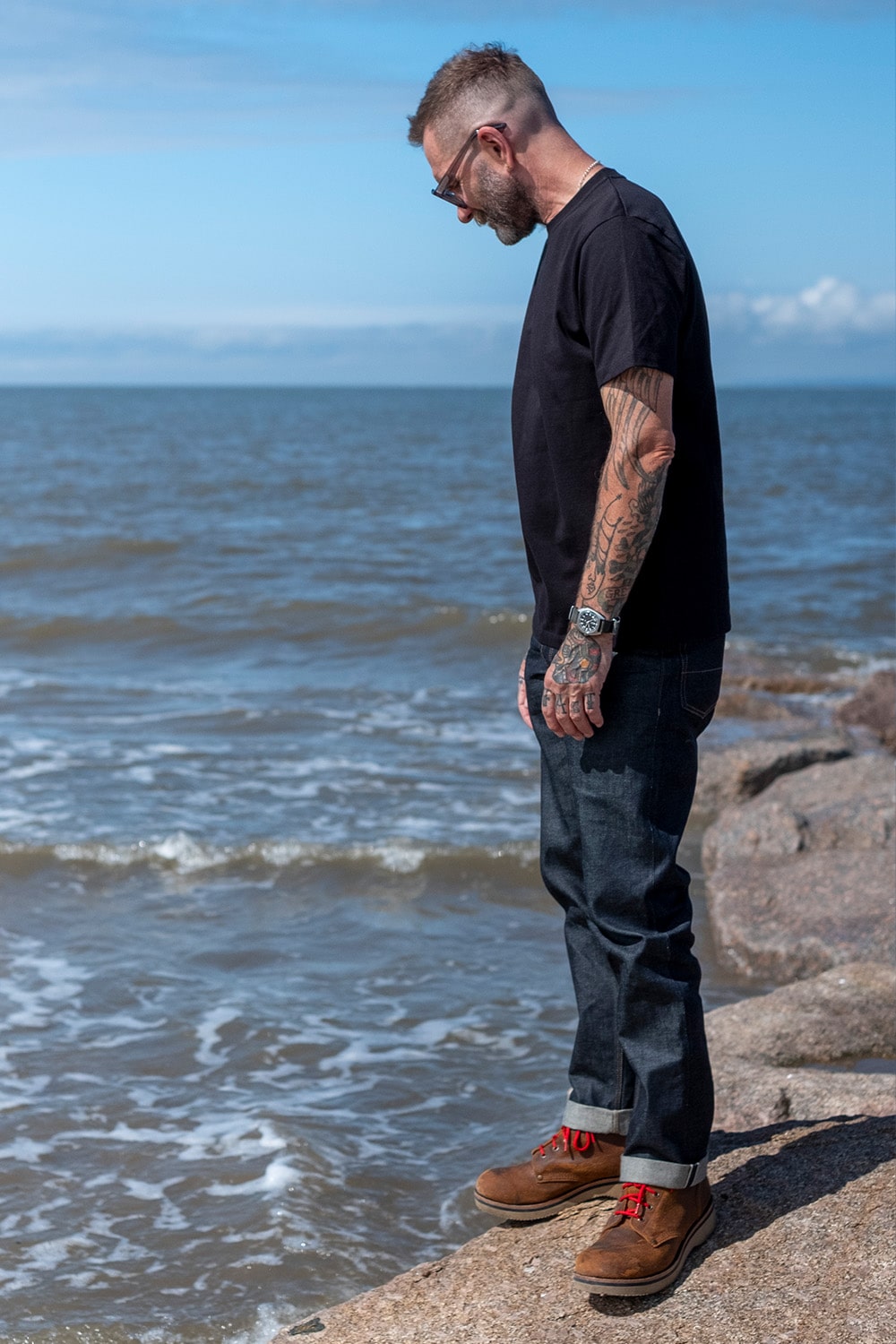 A man with tattoos stands on a rocky shoreline looking out at the ocean under a blue sky. He is wearing a black t-shirt, dark blue jeans, and brown boots with red laces.