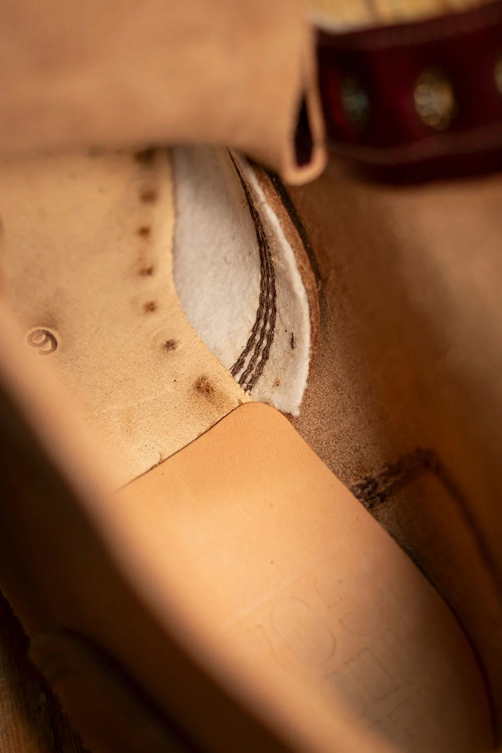 Close-up detail of a tan leather boot or shoe interior with visible stitching, a white insole section, and part of a dark reddish-brown strap or trim at the top.