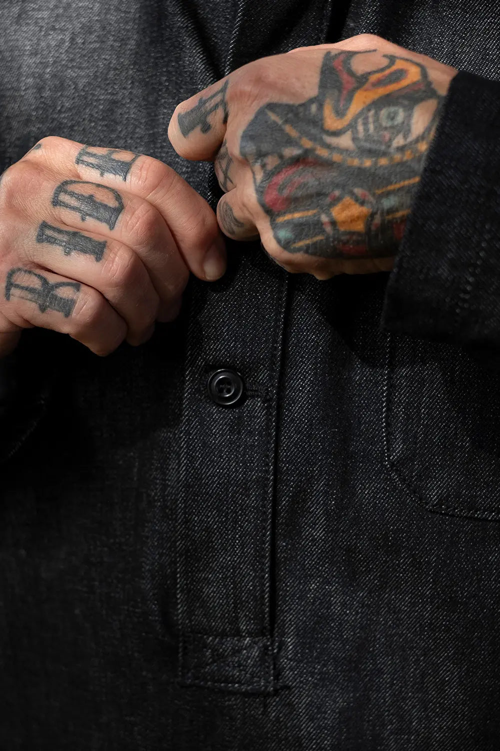 Close-up of a person's tattooed hands buttoning a dark charcoal gray textured overshirt or jacket.