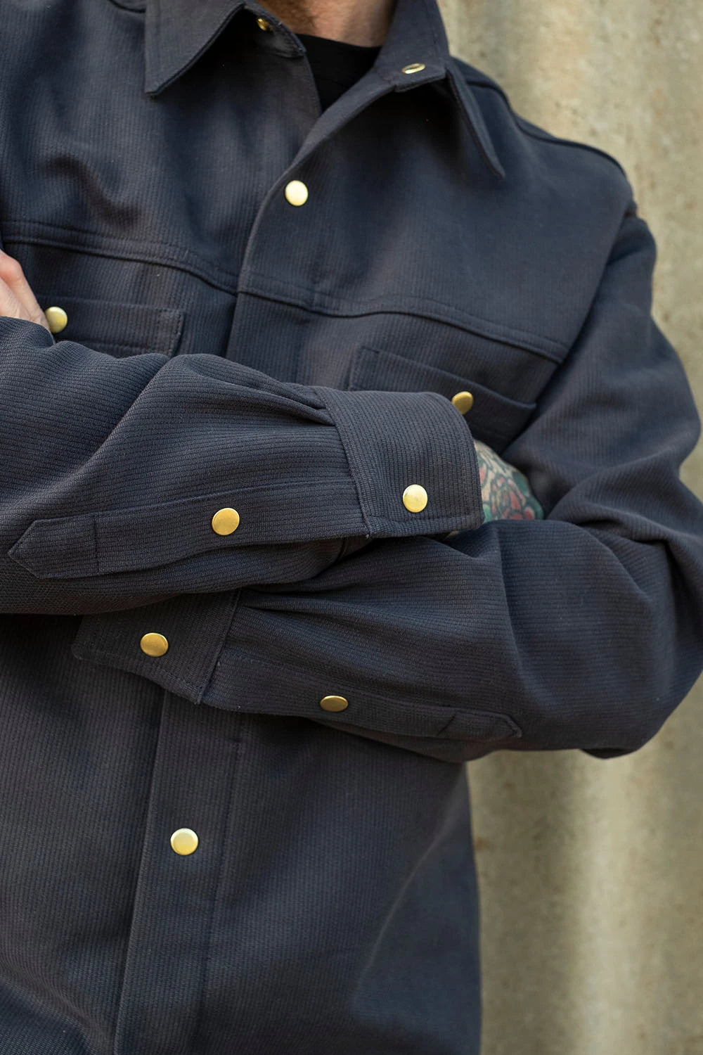 Close-up of a person wearing a dark navy bedford cord iron shirt with gold-tone snap buttons, with arms crossed against a textured concrete wall.