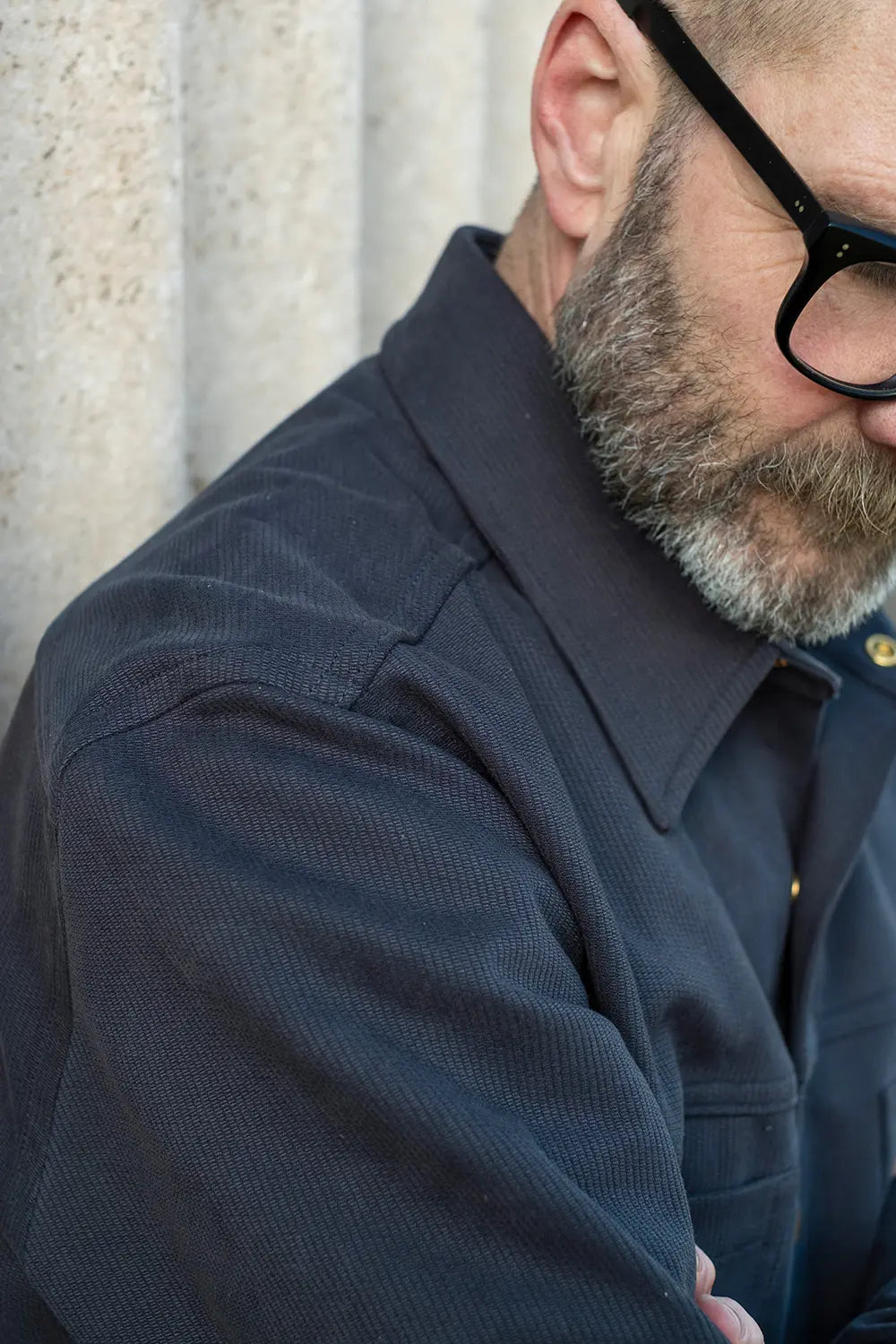 A close-up shot of Ed wearing a navy bedford cord ironshirt textured button-up shirt and black-framed glasses, leaning against a light stone wall.