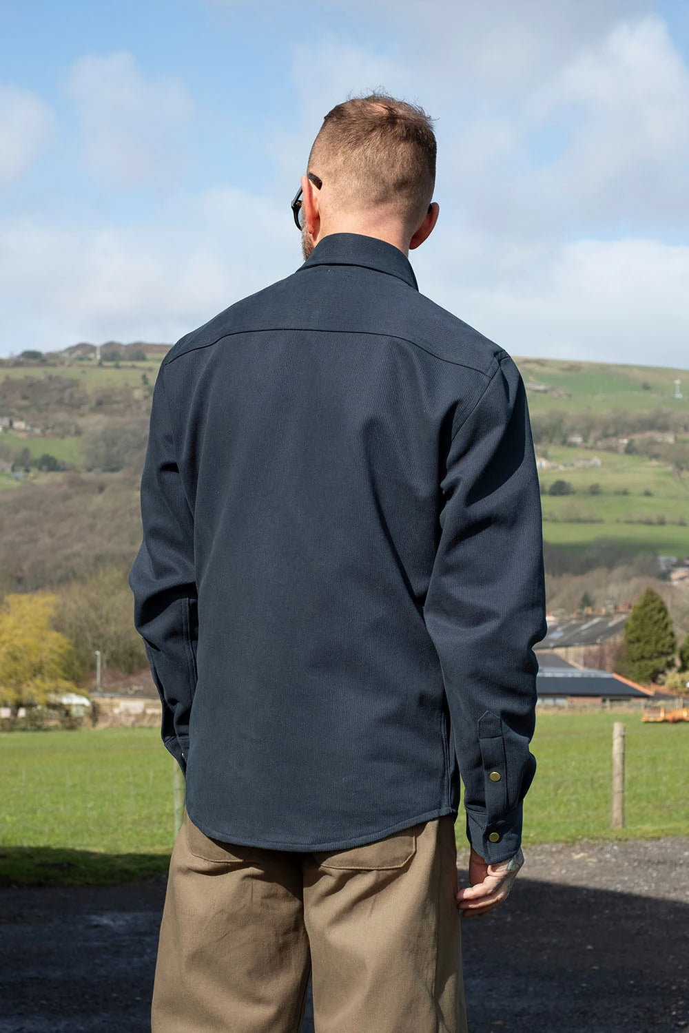 A man viewed from behind wearing a dark navy bedford cord iron shirt and khaki trousers, standing outdoors in a rural landscape with green hills and a blue sky.