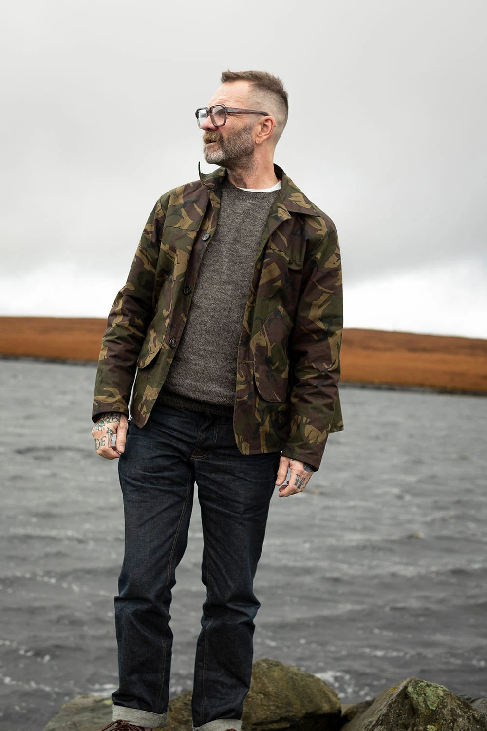 Ed wearing a green camouflage jacksnipe jacket over a gray knit sweater, dark jeans, and glasses stands on rocks by the water under an overcast sky.