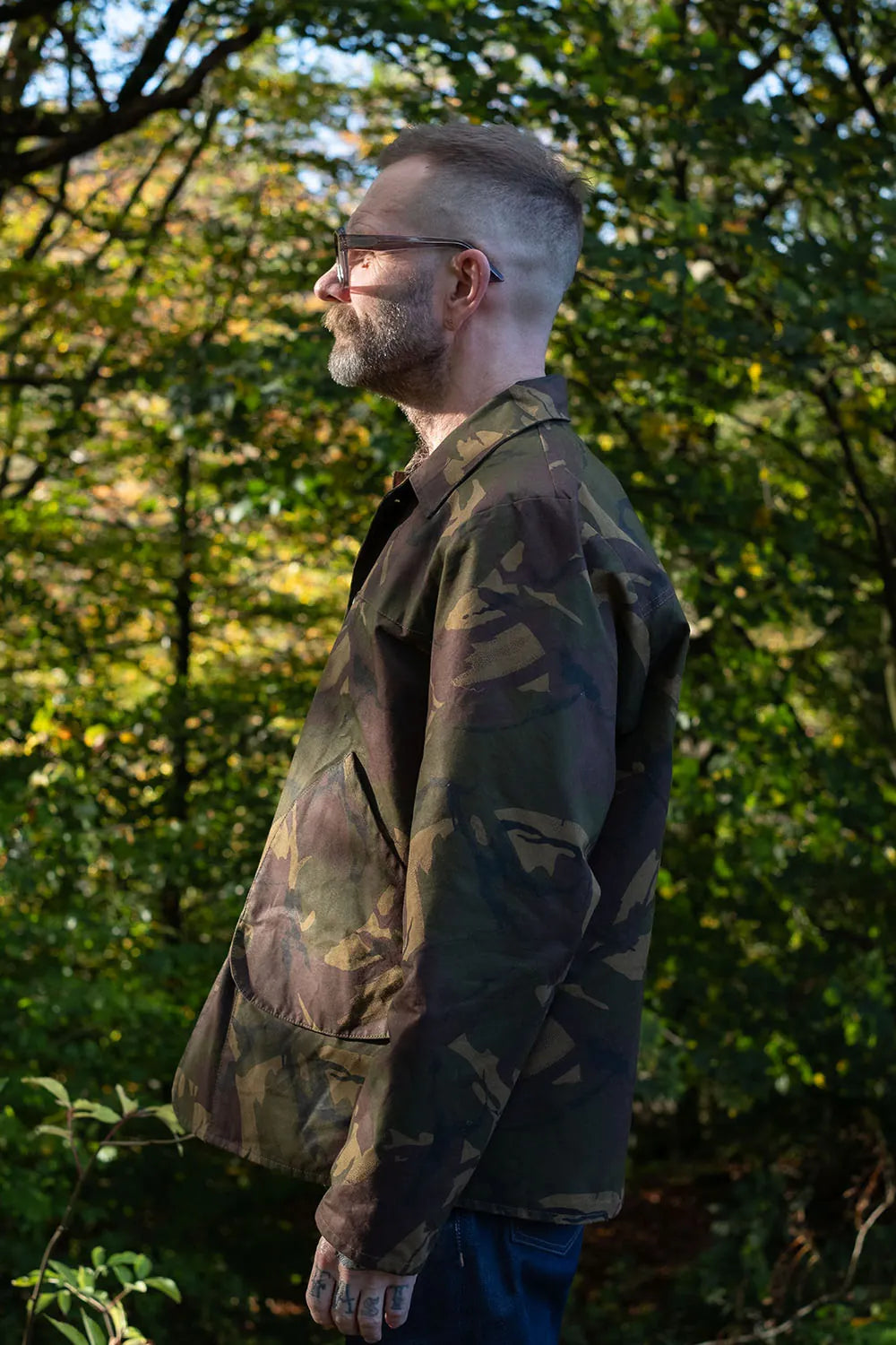 Ed in profile wearing a camouflage jacksnipe jacket and glasses, standing outdoors in a wooded area with green foliage.