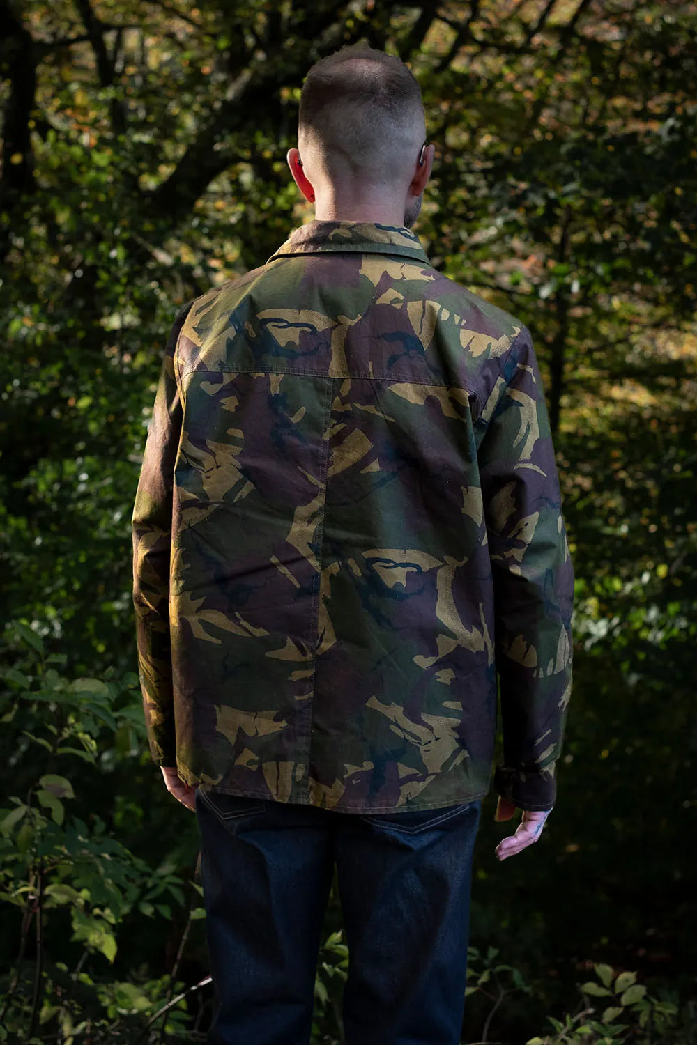 Ed viewed from behind wearing a camouflage jacksnipe jacket and dark blue jeans, standing in a dense green forest.