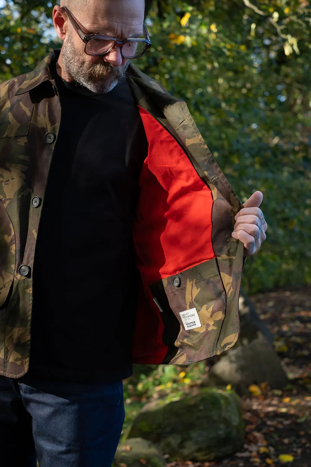 Ed outdoors wearing a camouflage jacksnipe jacket with a bright red interior lining, holding the jacket open to show the lining. He is also wearing a black shirt and blue jeans, with trees and rocks in the background.
