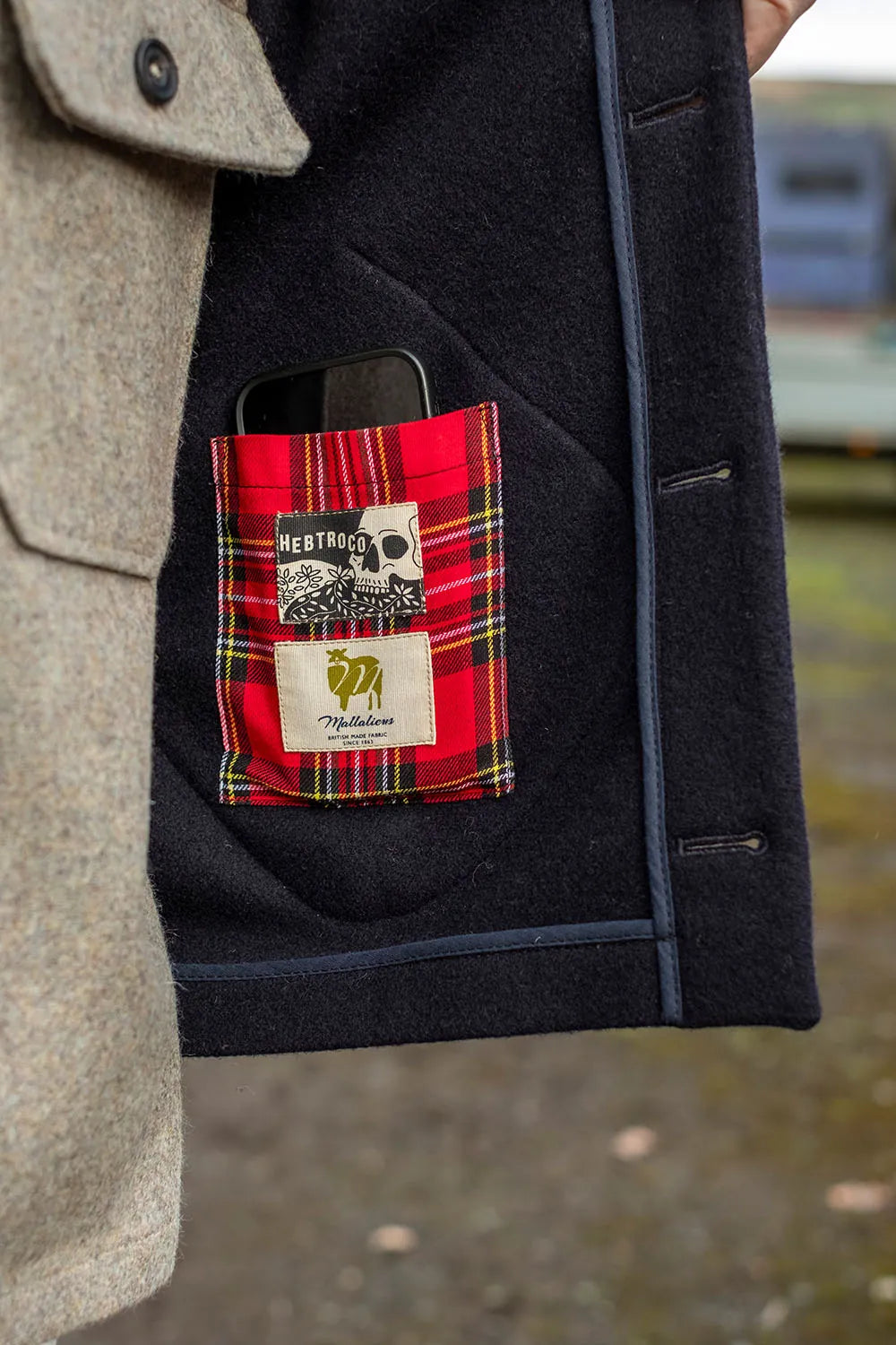 Close-up of a dark navy wool vest pocket holding a red tartan-patterned pocket, which contains a phone and two patches from Bertram's.
