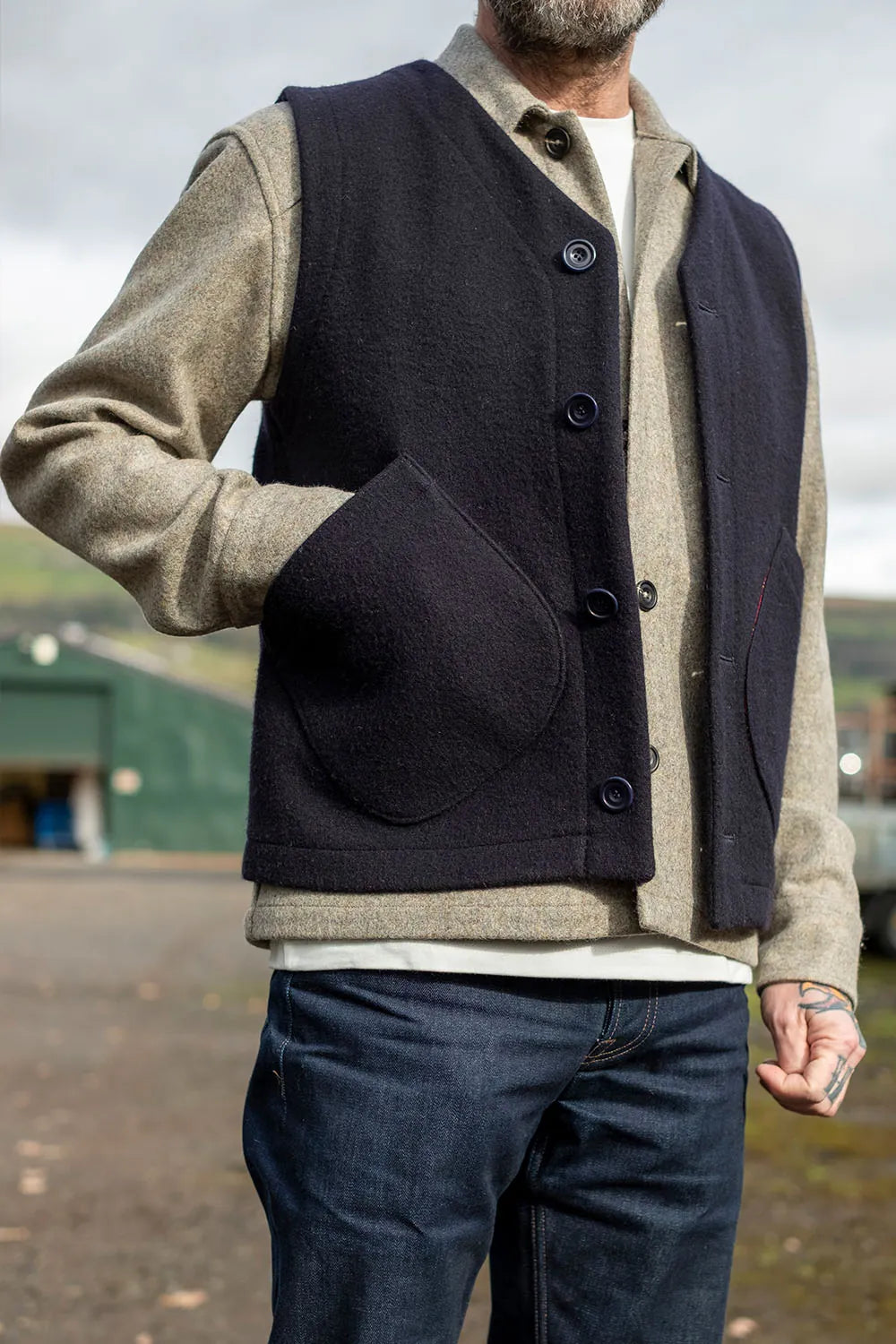 A man wearing a dark navy wool vest over a beige tweed shirt jacket, layered over a white t-shirt and paired with dark blue jeans, photographed outdoors.
