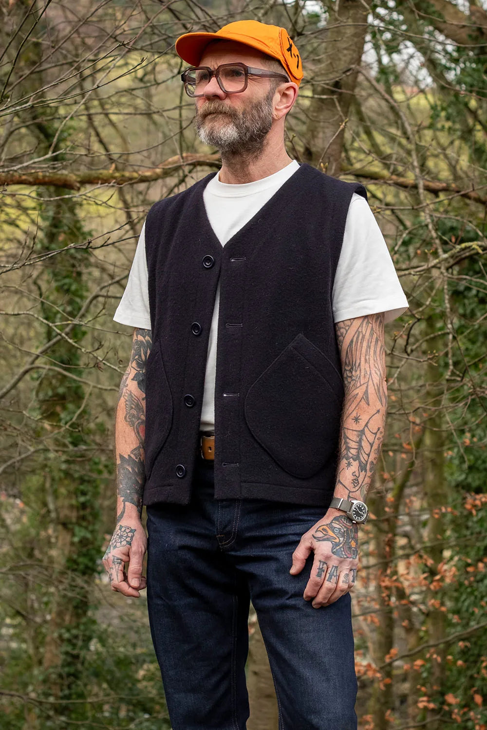 A man wearing a navy button-front wool vest over a white t-shirt, dark jeans, and an orange cap, standing outdoors in a wooded setting.