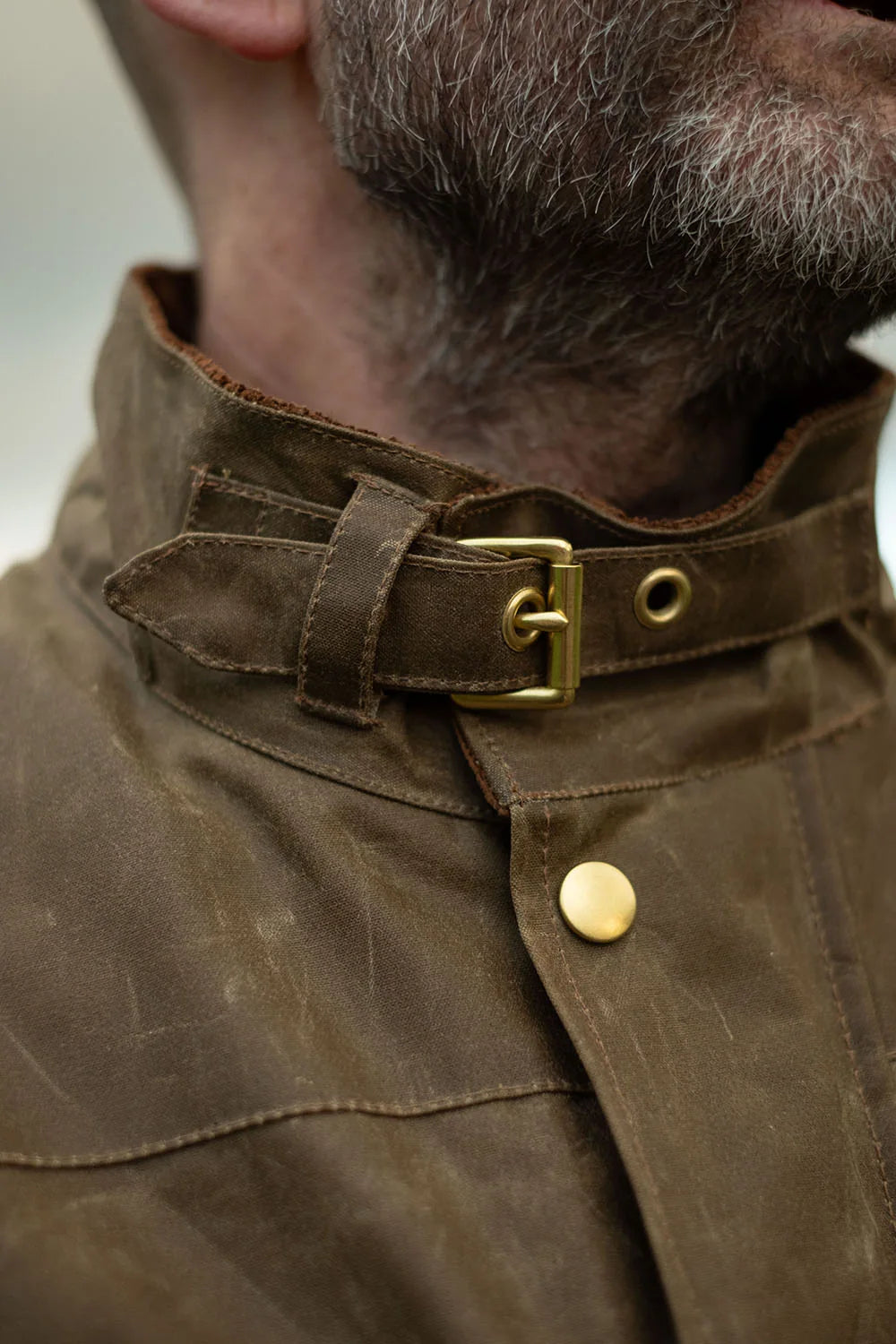 Close-up of a person wearing a dark brown waxed canvas jacket with a high collar, brass buckle strap, and brass snap button.