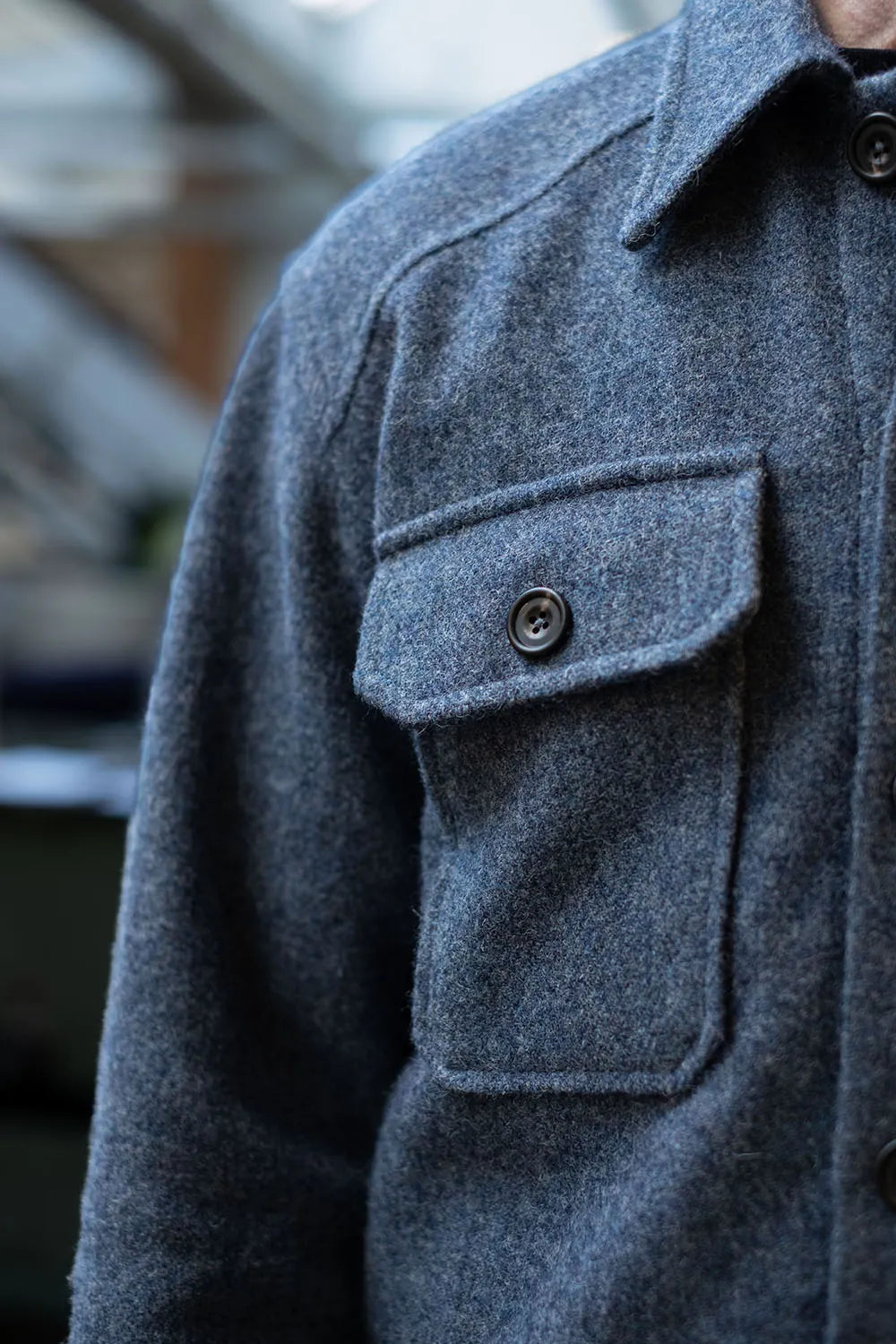 Close-up of a person wearing a USB36 Wool overshirt with a chest pocket and dark button, photographed outdoors with a blurred background.