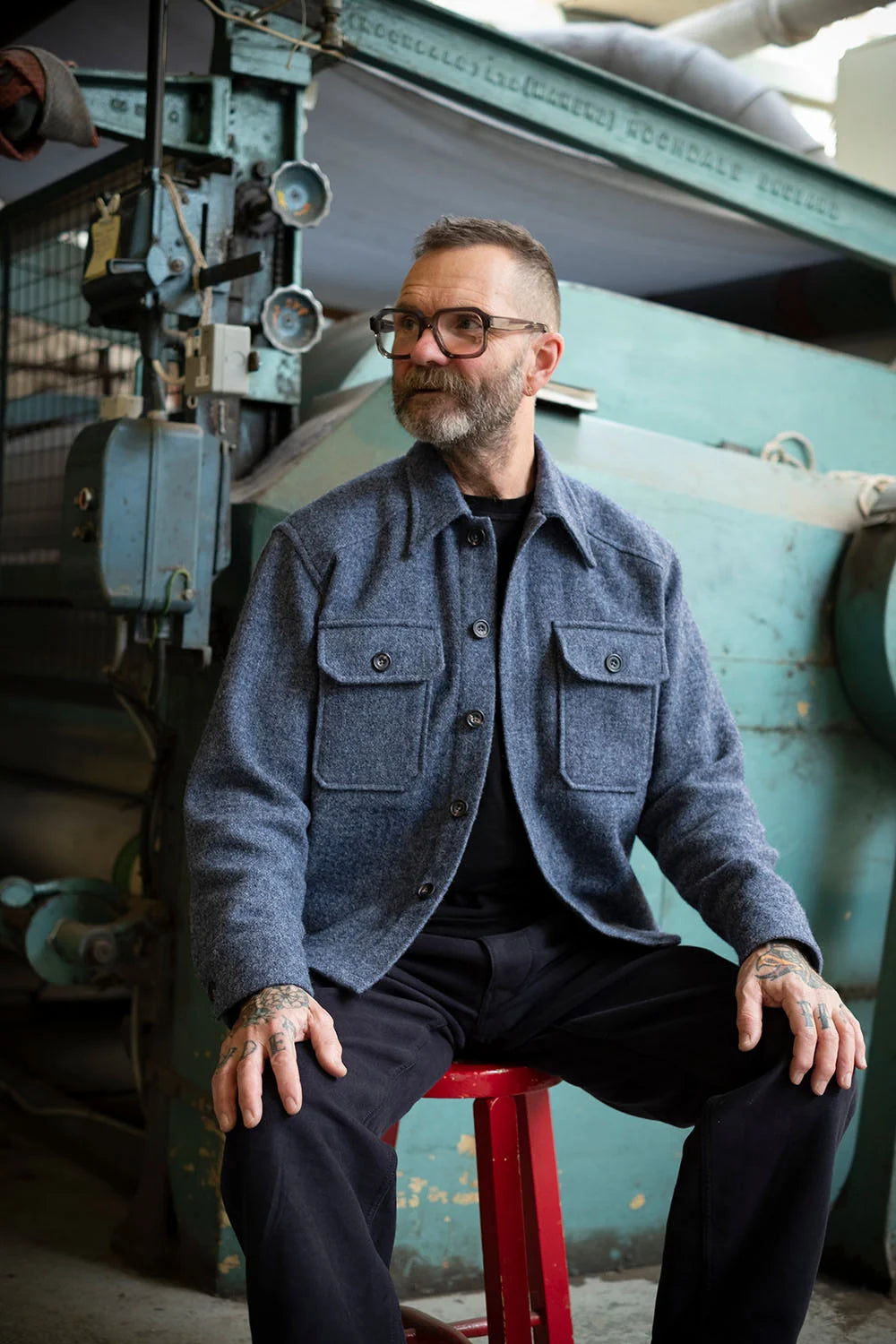 A man with glasses and a beard sits on a red stool in front of large vintage industrial machinery, wearing the USB36 blue-gray overshirt over a black shirt and black trousers.