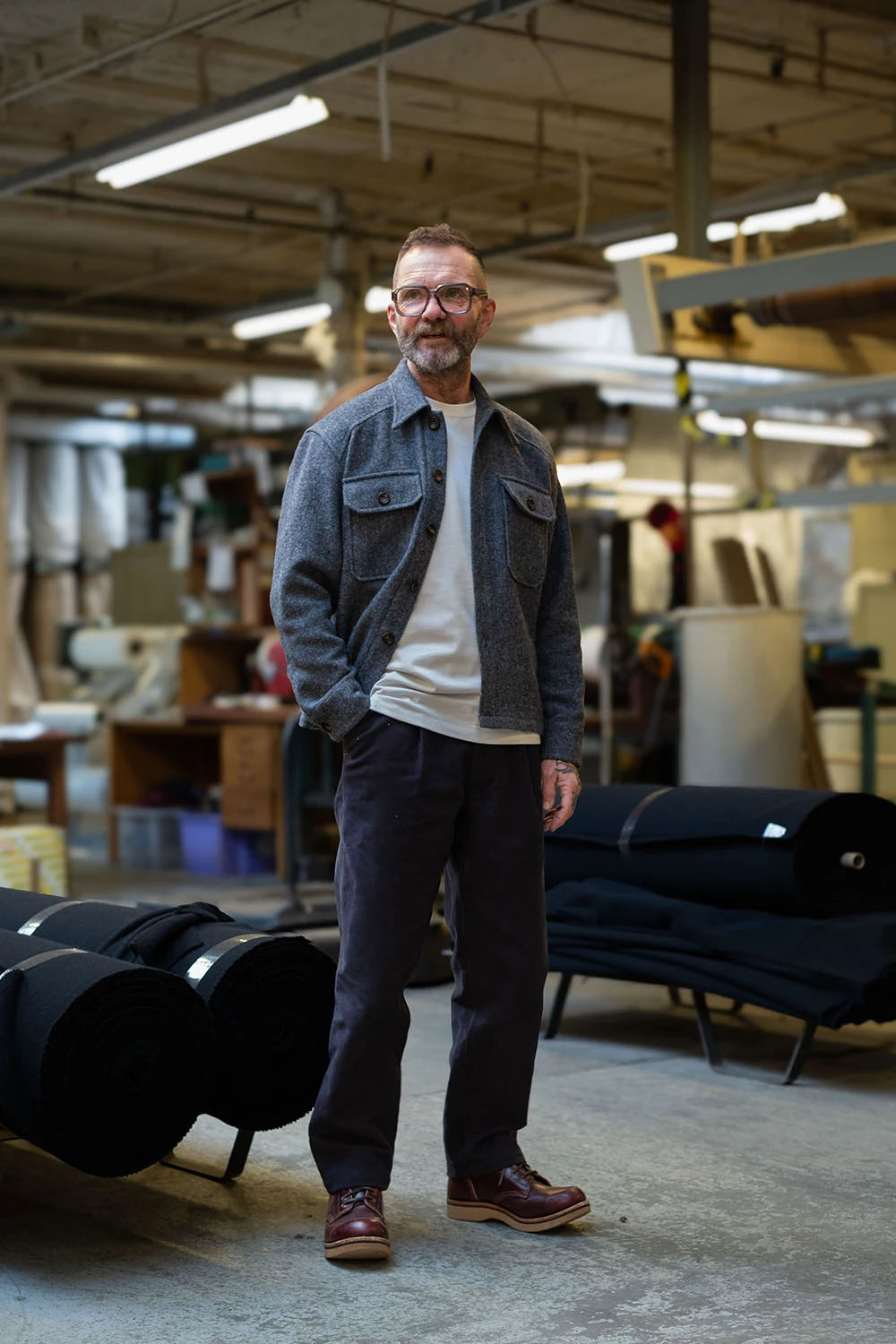 Ed wearing the USB36 wool overshirt over a white t-shirt, dark charcoal trousers, and brown leather shoes stands in a workshop environment.