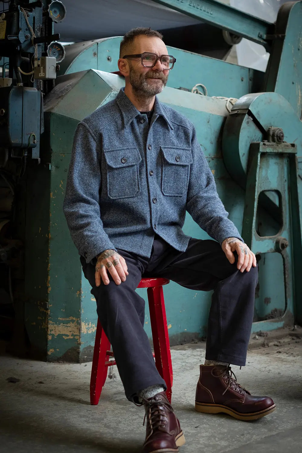 A man wearing a textured blue-gray button-up shirt jacket, dark trousers, and brown leather boots sits on a red stool in front of a large teal industrial machine.