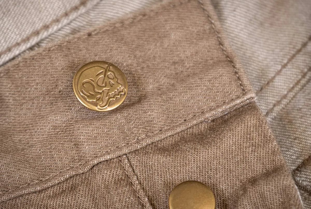 Close-up of a beige canvas garment detail featuring a round brass button with an embossed animal head logo.