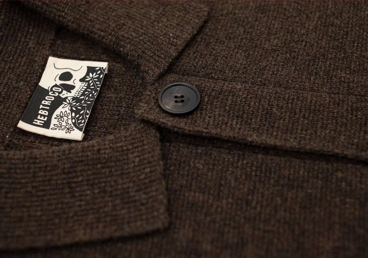Close-up of a dark brown textured wool jacket with a black button and a white sewn-in label featuring a black skull graphic and the brand name 'HEFT TOY'.