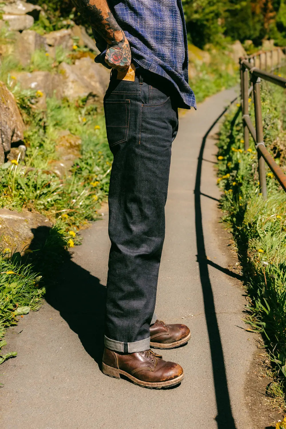 A person standing on a paved outdoor path wearing dark indigo denim jeans, a blue plaid shirt, and brown leather boots, with grass, rocks, and a wooden railing visible in the background.
