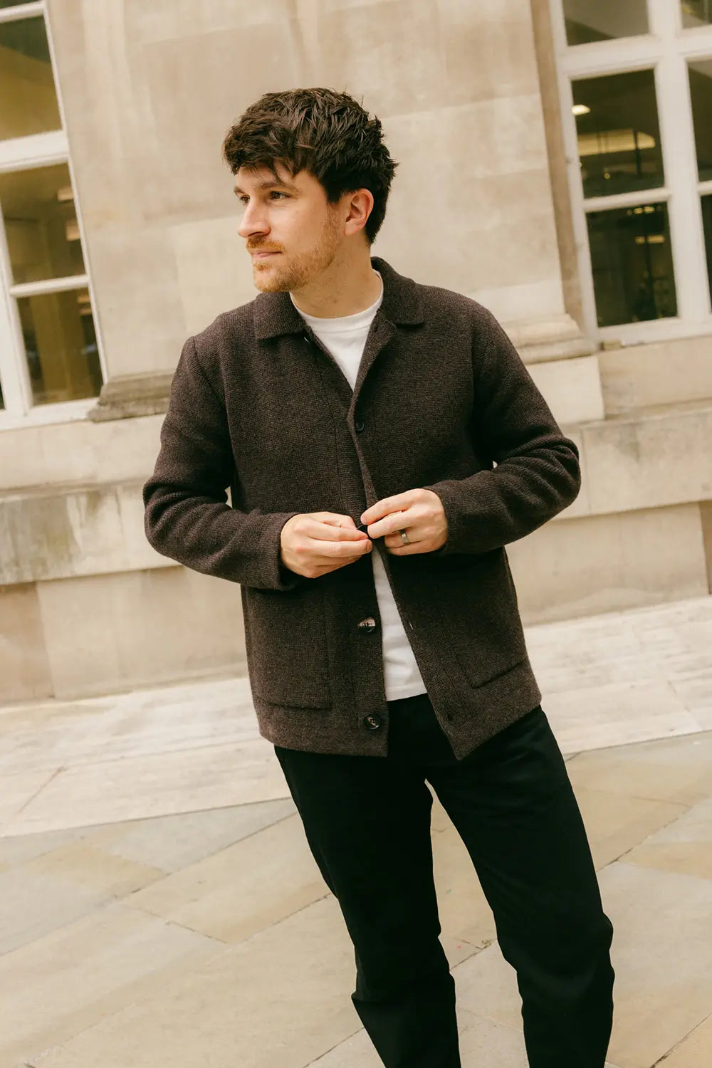 A man wearing a dark brown wool-blend cardigan over a white t-shirt and black trousers, standing outdoors in front of a stone building.